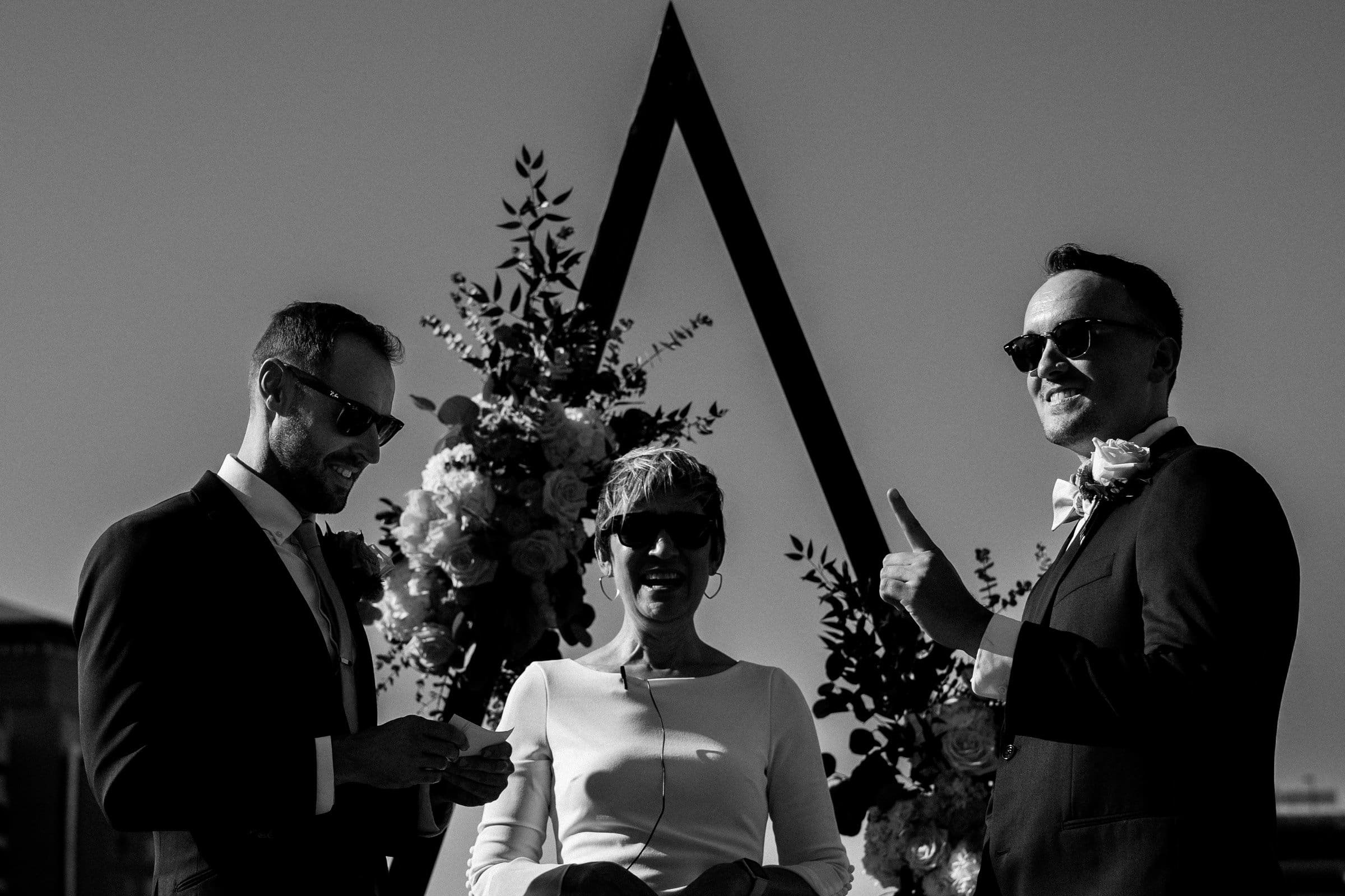 A black and white photo captures a joyful moment at an outdoor wedding ceremony. Two men in suits and sunglasses flank a woman in a dress under a geometric floral arch. Their smiles mirror the elegance seen in Union Station Raleigh wedding photography.