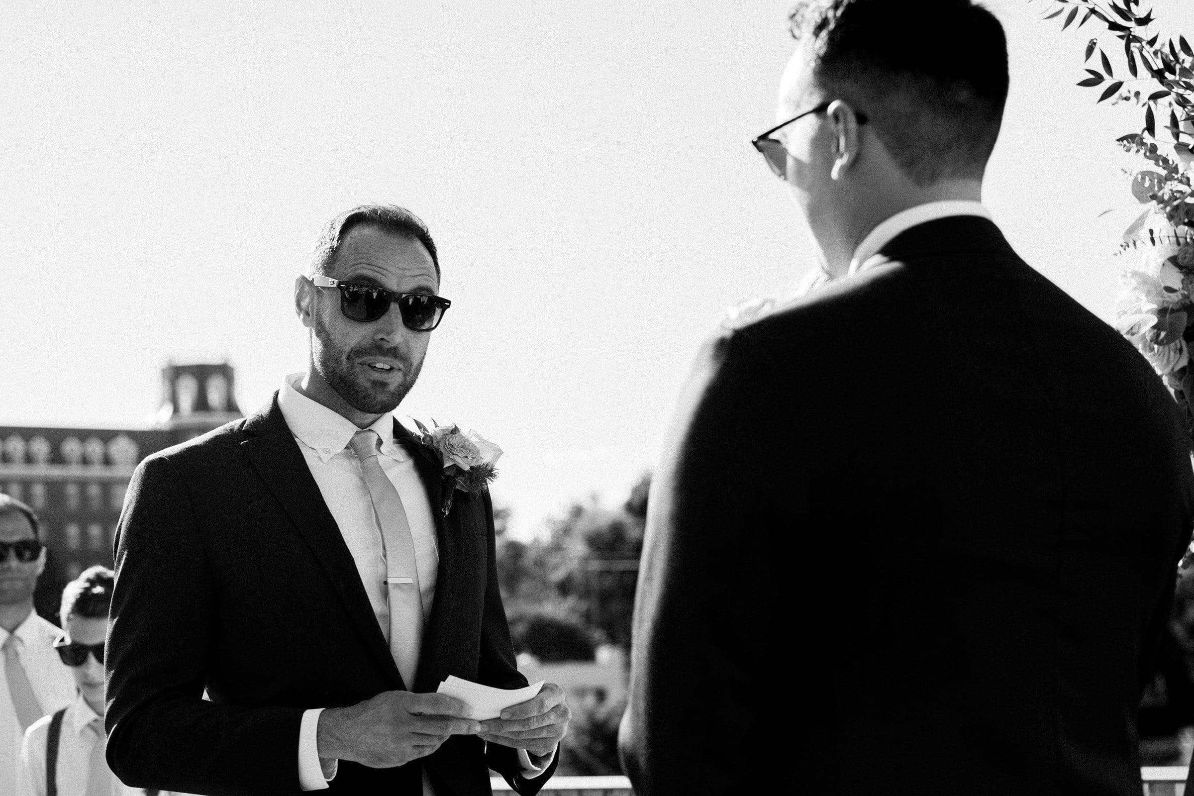 A man in a suit and sunglasses reads vows to another man during an outdoor ceremony, captured in timeless black and white. Guests are seated in the background, witnessing this special moment at Union Station Raleigh.
