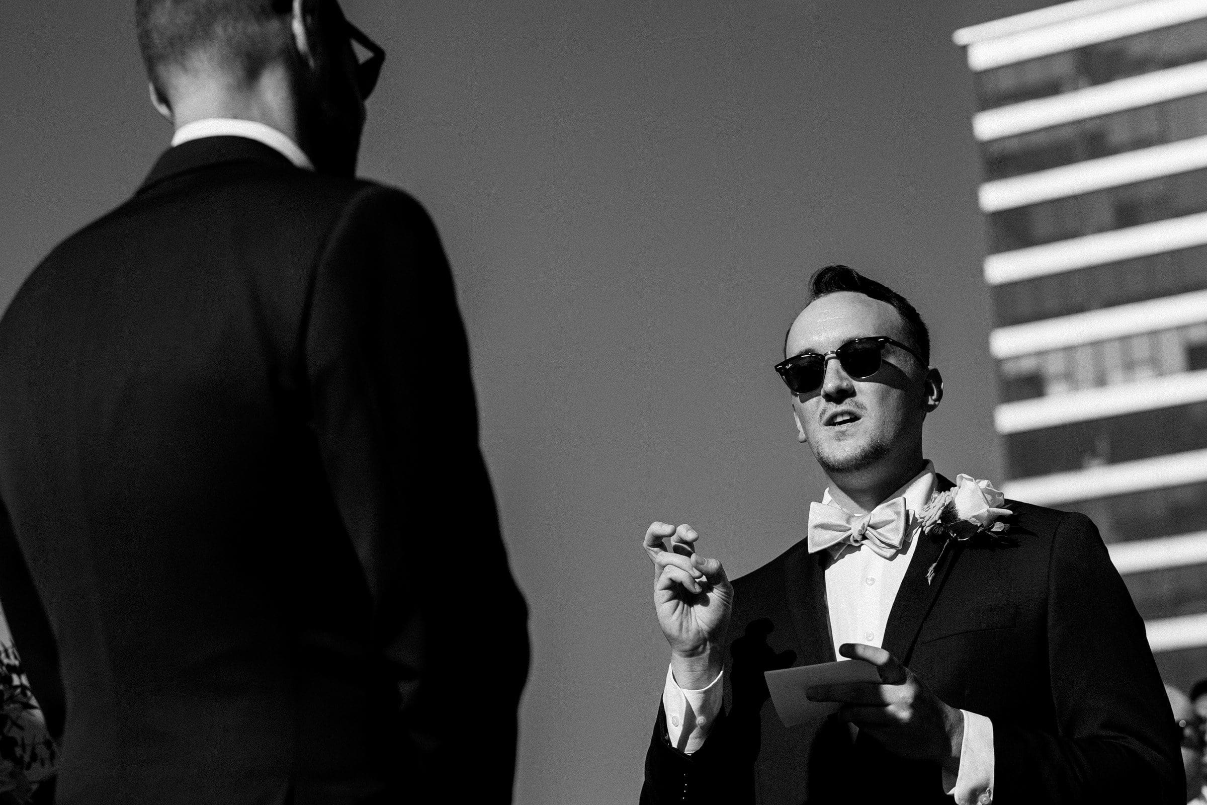 A black-and-white photo captures two people in suits and sunglasses at an outdoor event. One person holds a piece of paper, gesturing as if speaking. A tall building, reminiscent of Union Station, looms in the background, adding a touch of elegance akin to Raleigh wedding photography.