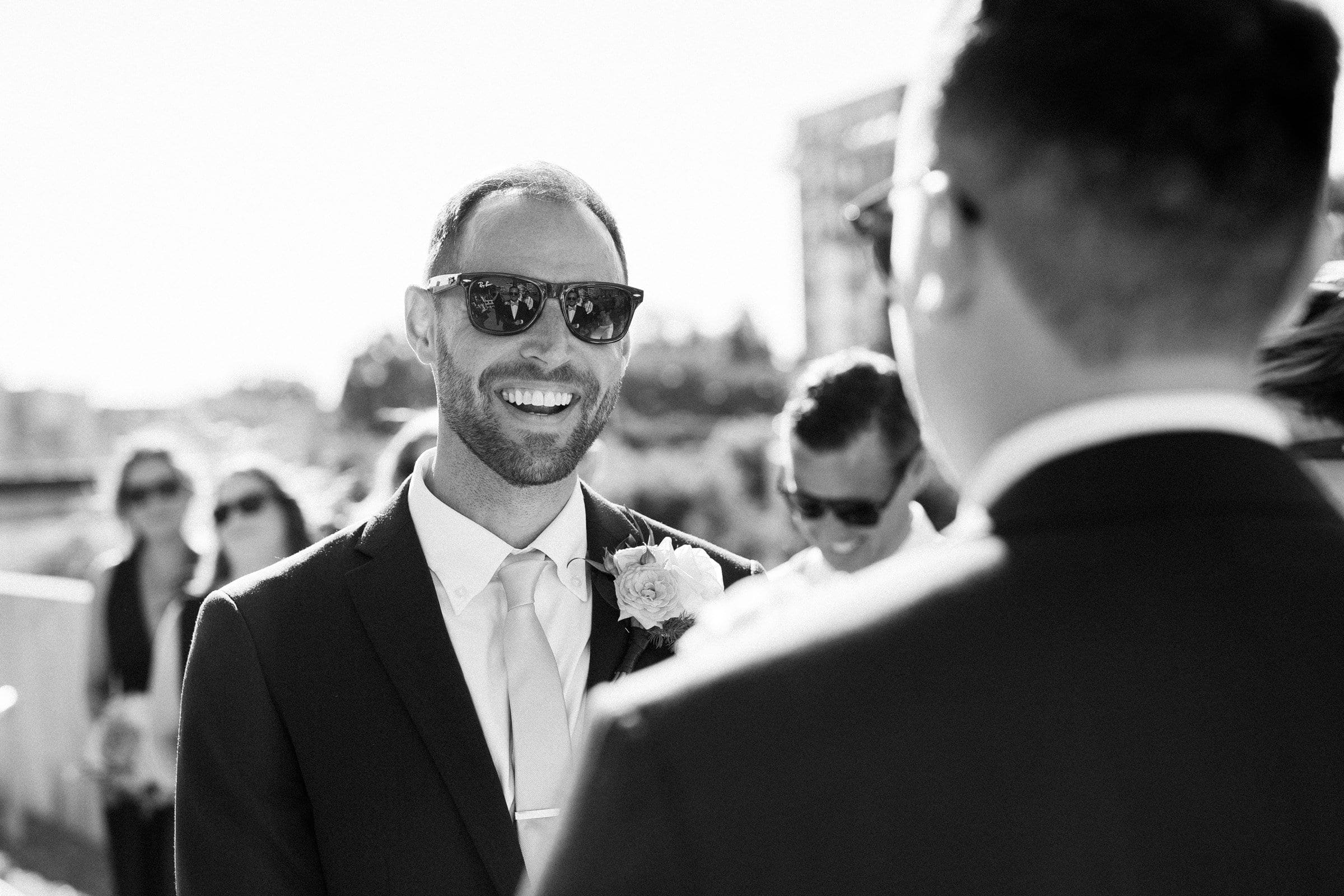A black and white photo captures a smiling man in sunglasses and a suit with a rose boutonniere, engaged in conversation. The scene, reminiscent of timeless Raleigh wedding photography, features blurred figures and Union Station's architecture in the background.