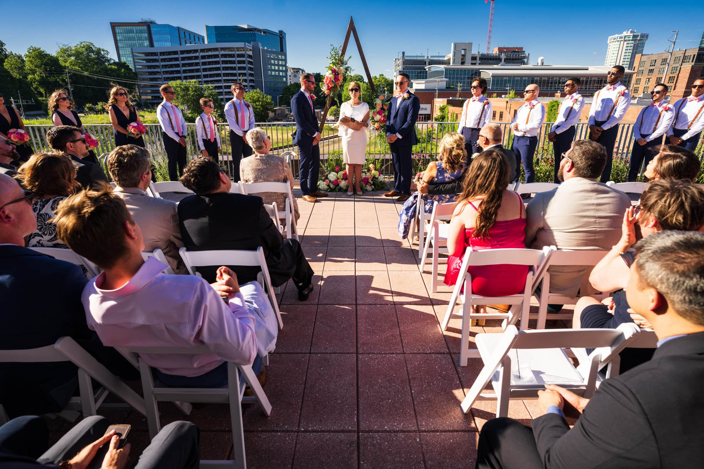 On a sunny day, the couple stands before a geometric arch at Union Station in Raleigh, surrounded by their wedding party. Guests enjoy the rooftop setting amidst cityscapes, capturing every moment with exquisite wedding photography.