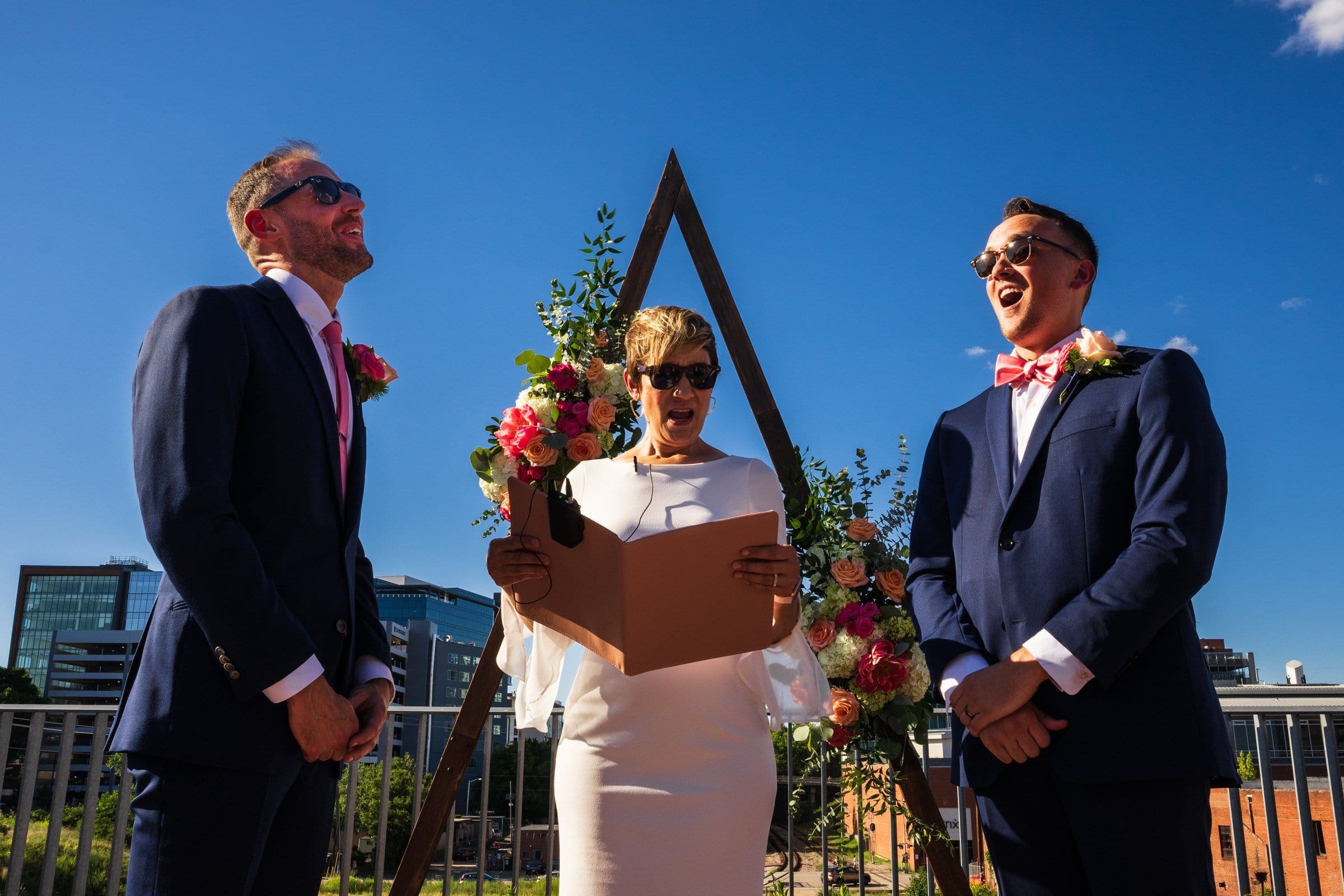 A wedding ceremony on a sunny day captures the joy of the moment, with two people in suits standing beside the officiant, all smiling and donning sunglasses. The scene is beautifully framed by a triangular wooden arch adorned with flowers, reminiscent of those stunning Union Station Raleigh wedding photographs.