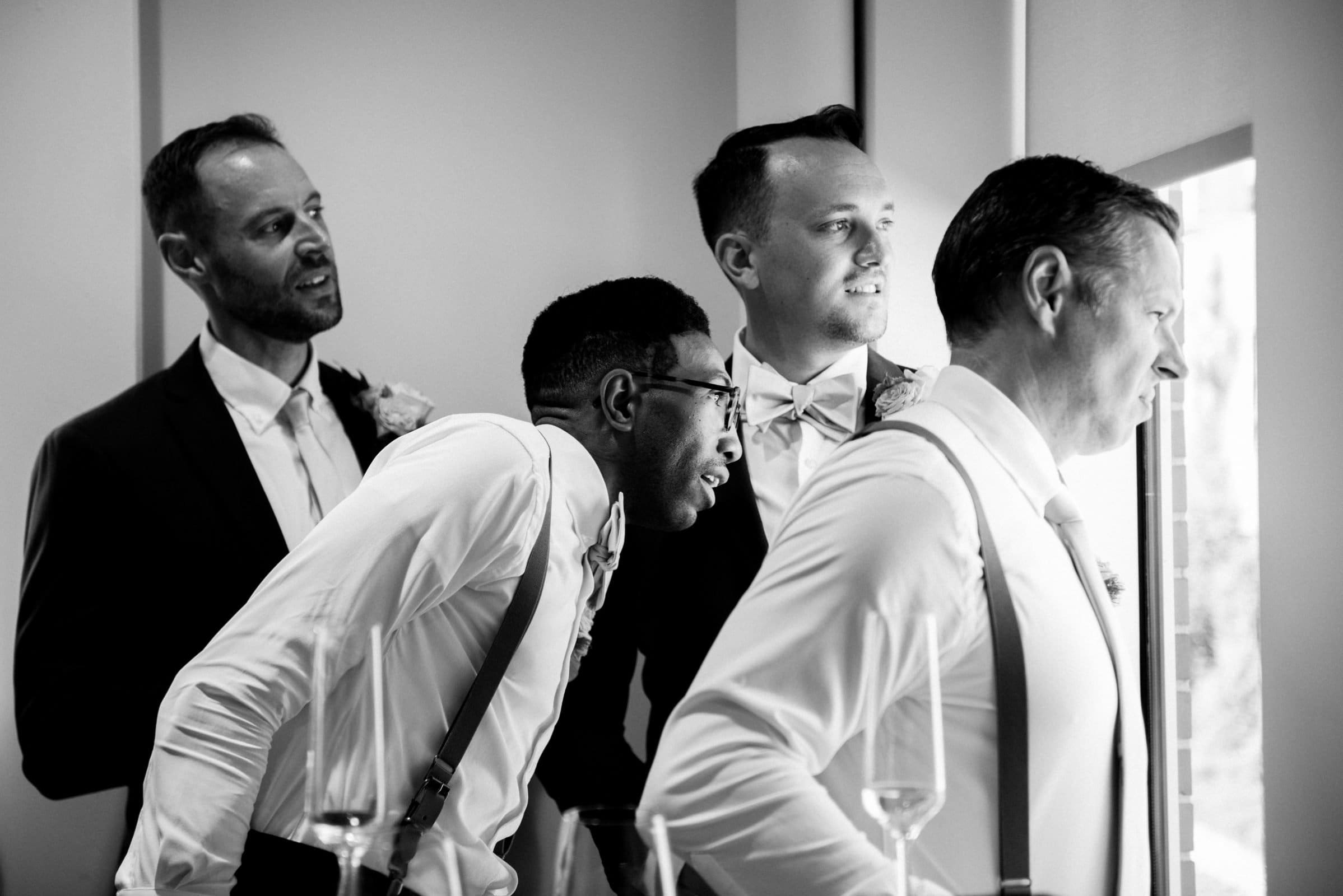 Four men in formal attire eagerly look out a window at Union Station Raleigh. Two wear suspenders, and all have boutonnieres. They appear excited, as if wedding photography captures this special moment in the well-lit room.
