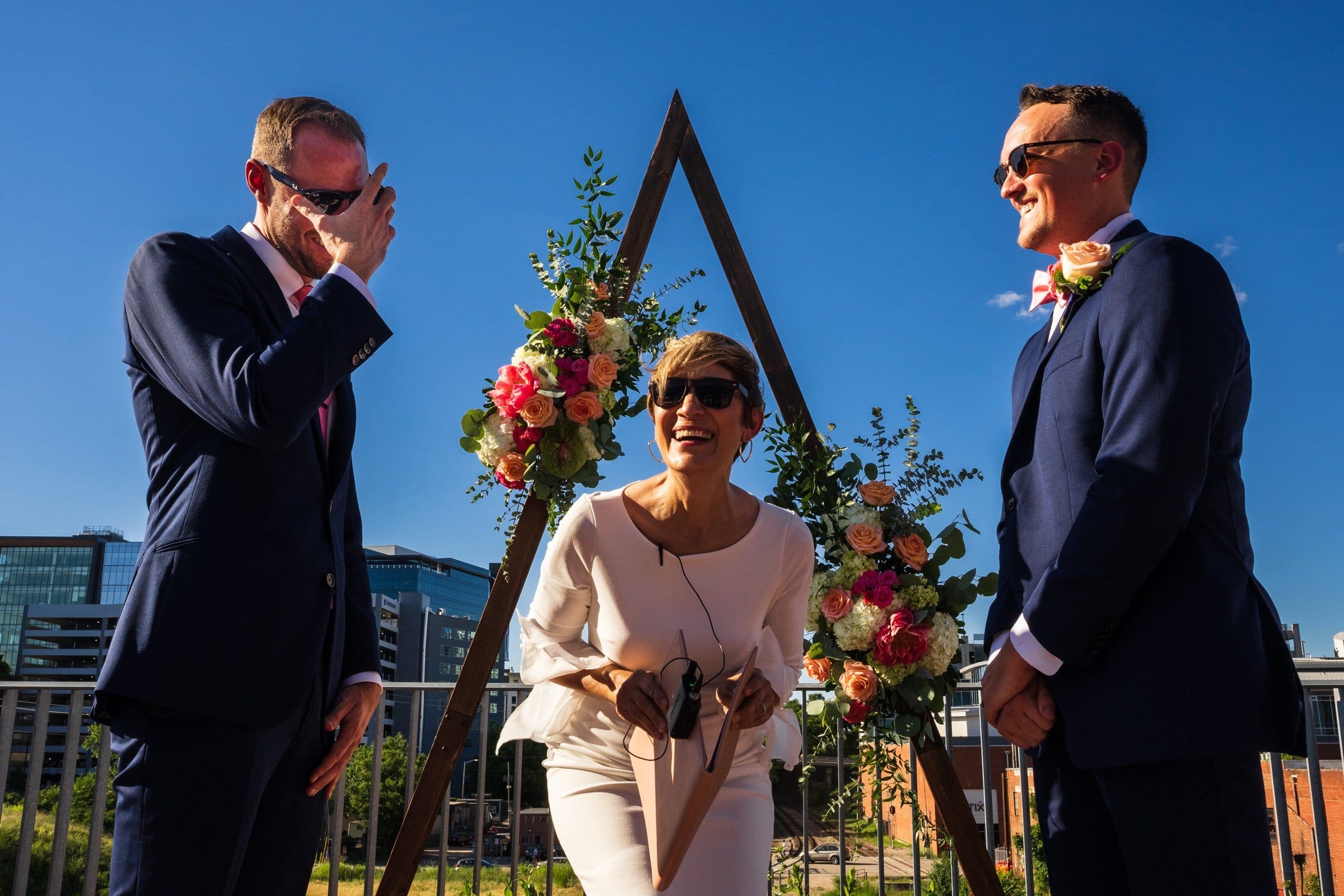 Three people are at an outdoor wedding ceremony. A woman in a white dress stands smiling under a floral arch, holding a microphone. Two men, dressed in suits, share a laugh. The blue sky and city buildings reminiscent of Union Station Raleigh create the perfect backdrop for stunning wedding photography.