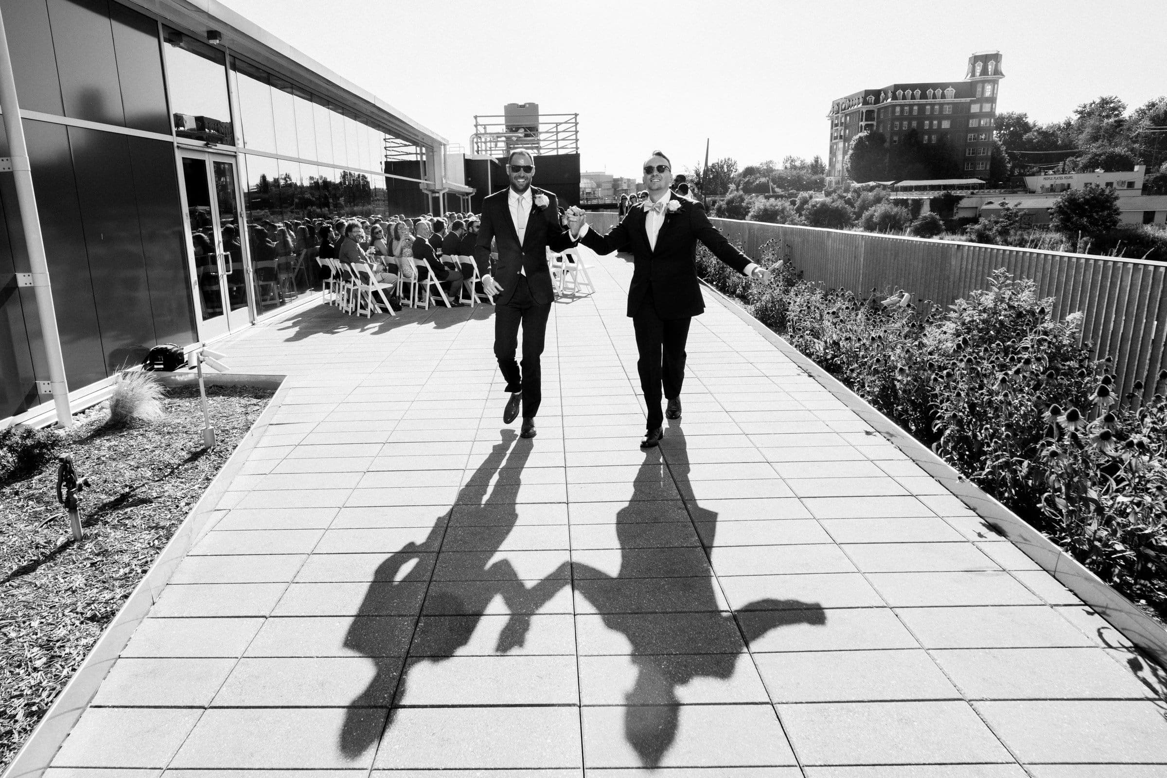 Two people in suits joyfully walk hand in hand down a sunlit path outside Union Station, their shadows cast behind them. A seated crowd is in the background near a modern building, and trees dot the horizon. The scene conveys celebration and happiness, captured perfectly by wedding photography.