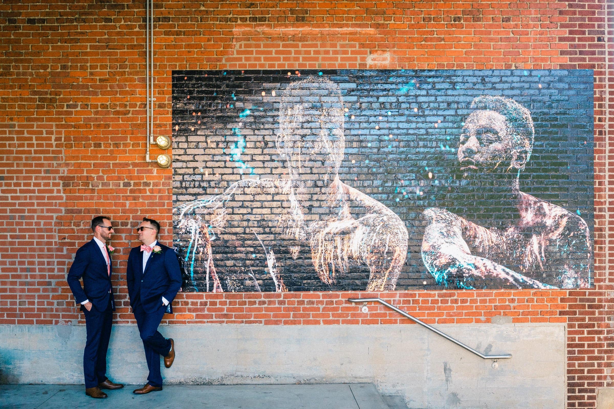 Two men in suits stand on a sidewalk against a brick wall, each leaning on one leg and facing each other. They are in front of a mural depicting two figures. Reminiscent of Union Station Raleigh's elegance, this scene captures the charm of candid wedding photography. A small metal railing is visible to the right.