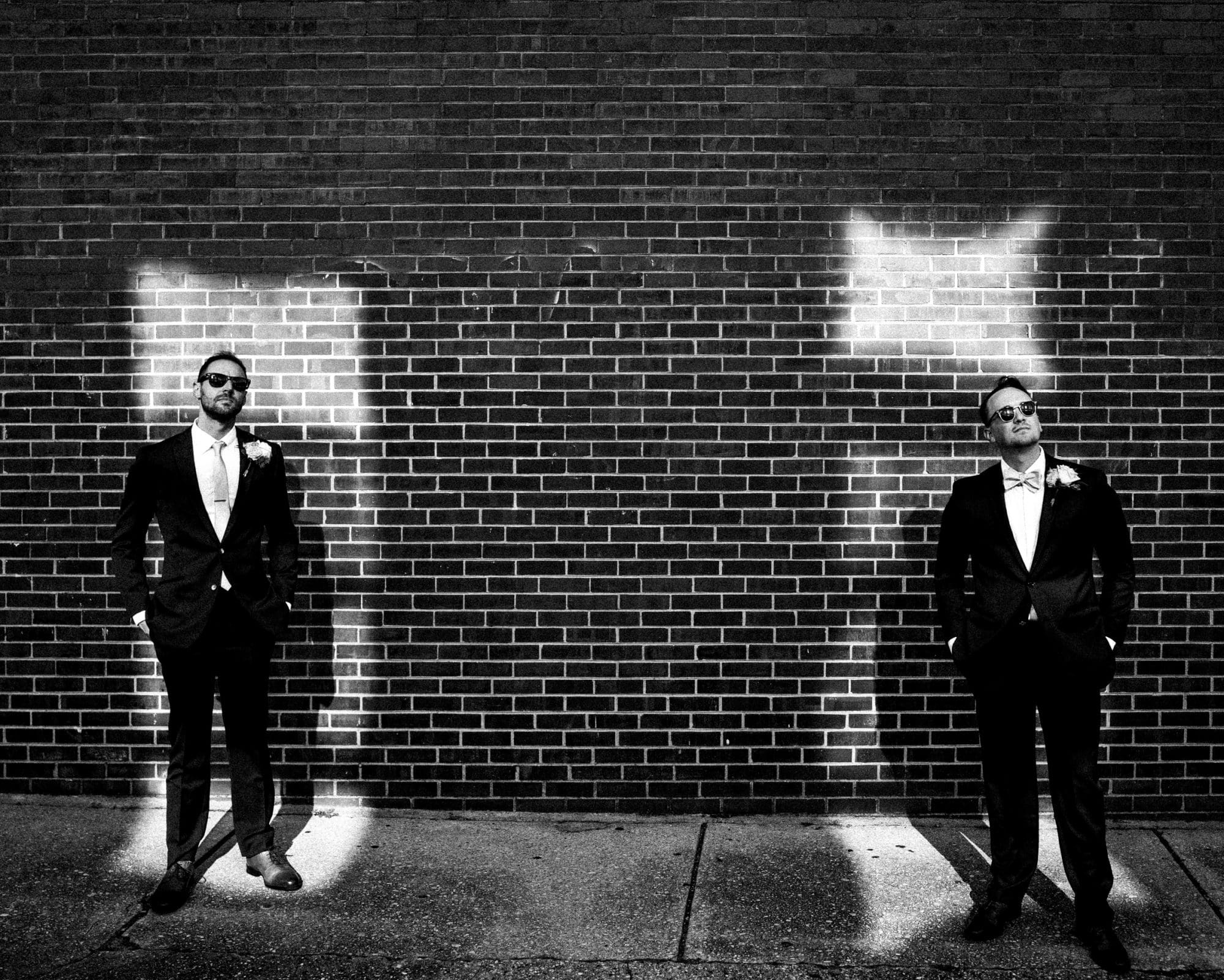Two men in suits stand apart in front of a brick wall with bright light patterns behind them at Union Station Raleigh. Both wear sunglasses and have hands in pockets, with corsages on their jackets, creating a dramatic, high-contrast black and white wedding photograph.