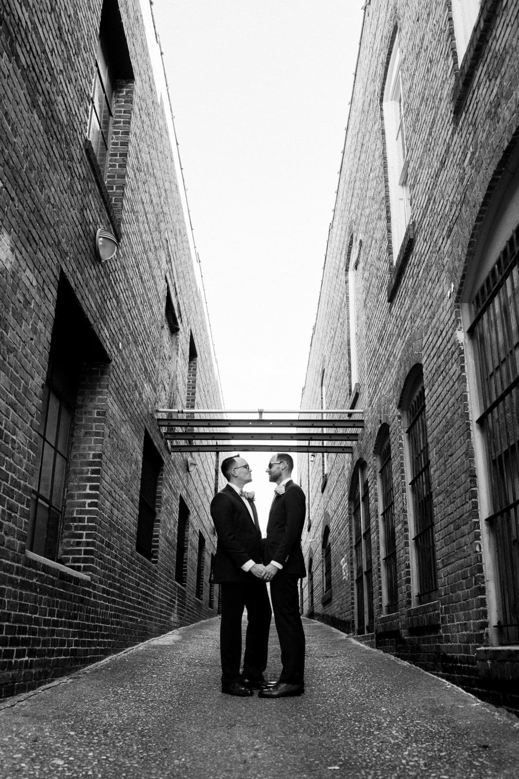 Two people in suits stand facing each other, holding hands, in a narrow alleyway between brick buildings. The perspective emphasizes the height of the structures and their symmetrical lines, reminiscent of classic wedding photography. The image is in black and white.