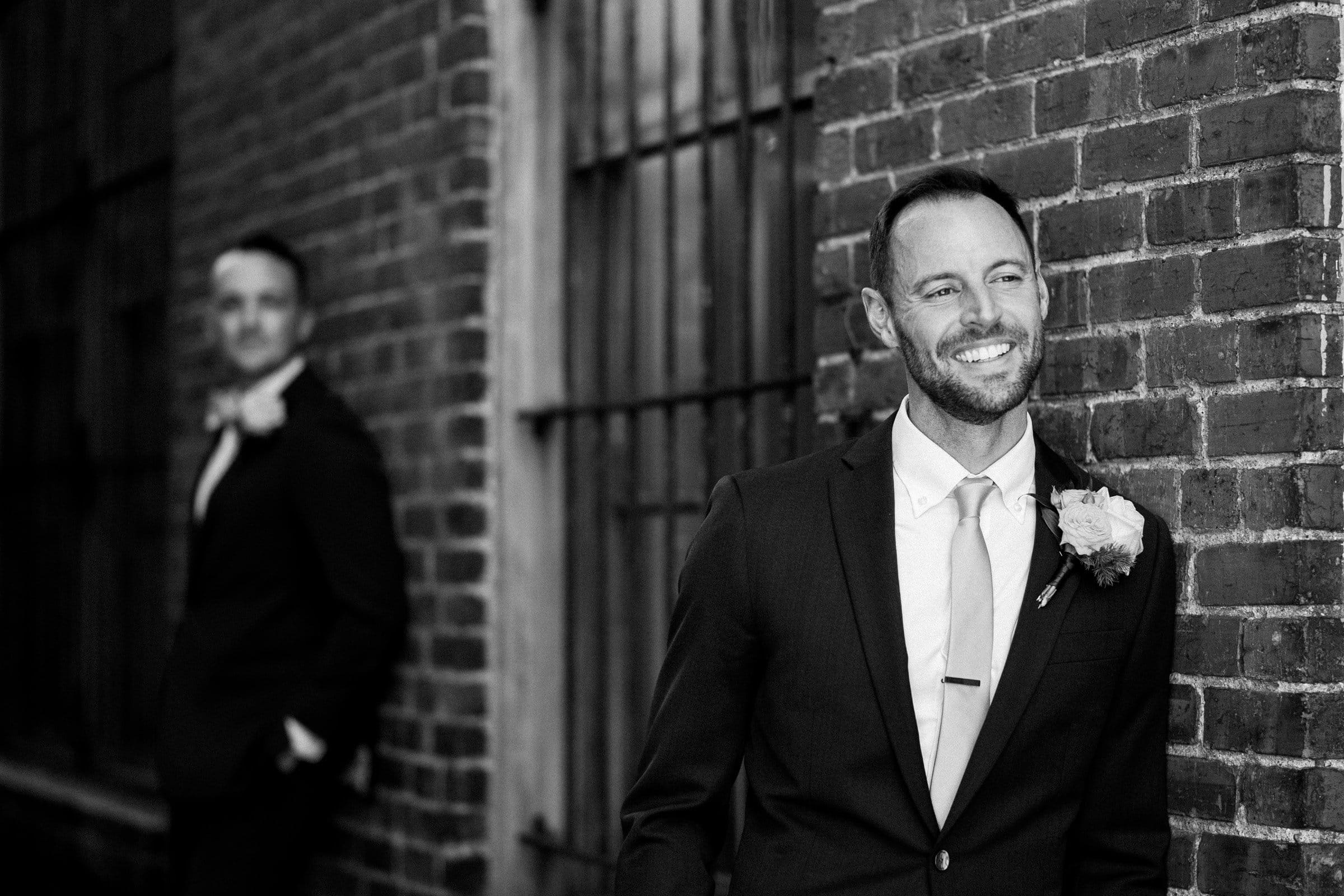A black and white photo captures two men in suits with boutonnières. One smiles, leaning against a brick wall, reminiscent of a classic Union Station Raleigh wedding photography moment, while the other stands in the background, partially visible and looking to the side.