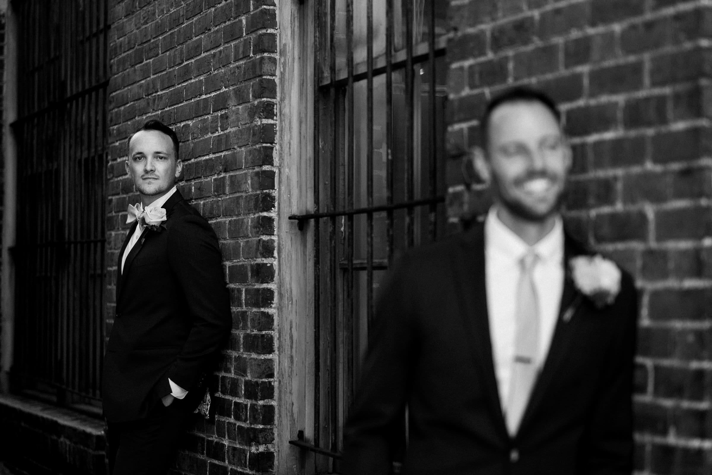 Two men in suits are standing against a brick wall. One, in focus on the left with hands in pockets, looks steady. The other is blurred in the foreground, smiling. Both have boutonnieres on their jackets—a perfect capture of style and joy reminiscent of a Union Station Raleigh wedding photography session.
