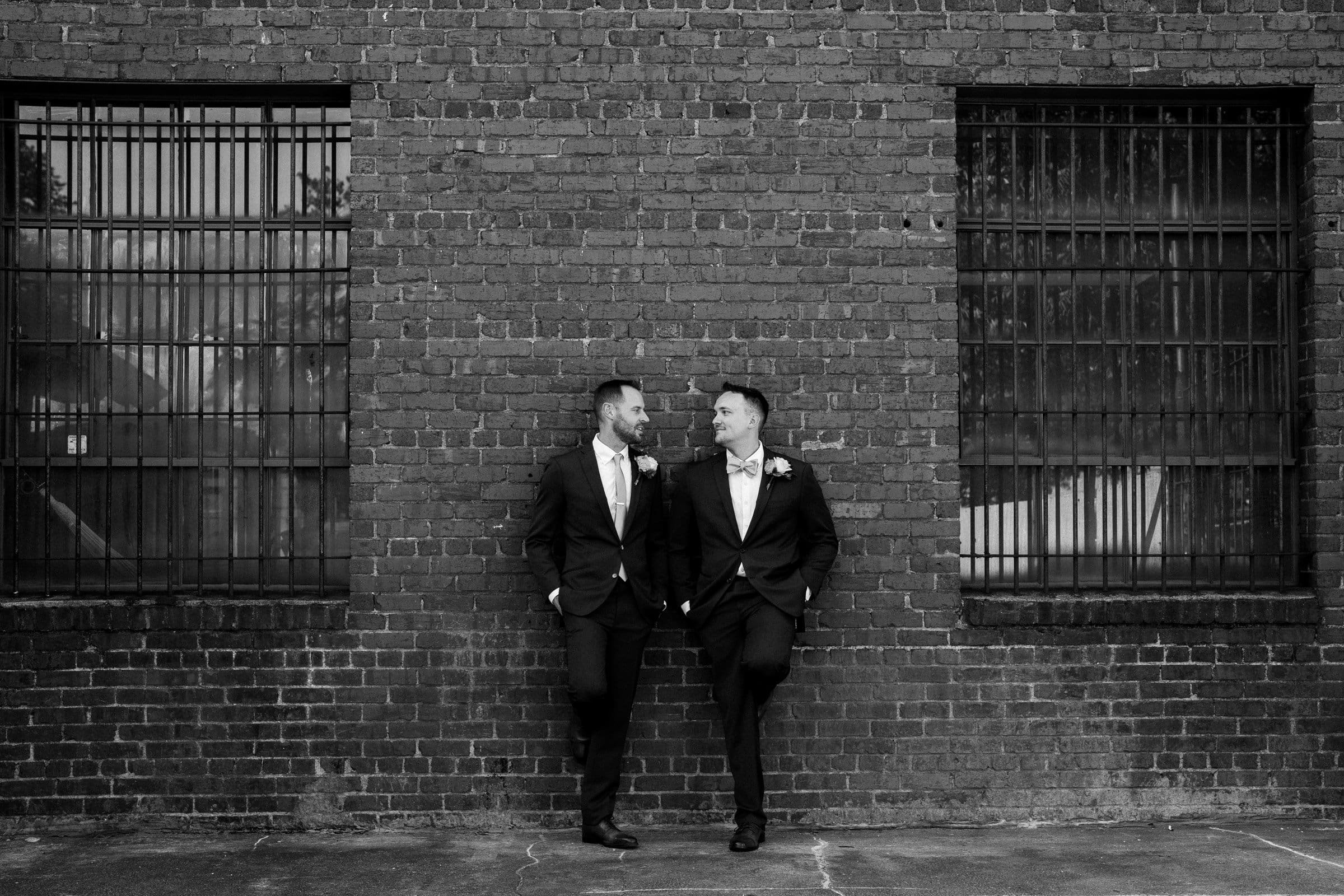 Two people in suits stand casually against a brick wall with barred windows, smiling at each other. With boutonnieres hinting at a formal occasion, like a wedding or celebration, this elegant black and white shot perfectly captures the charm of Union Station Raleigh wedding photography.