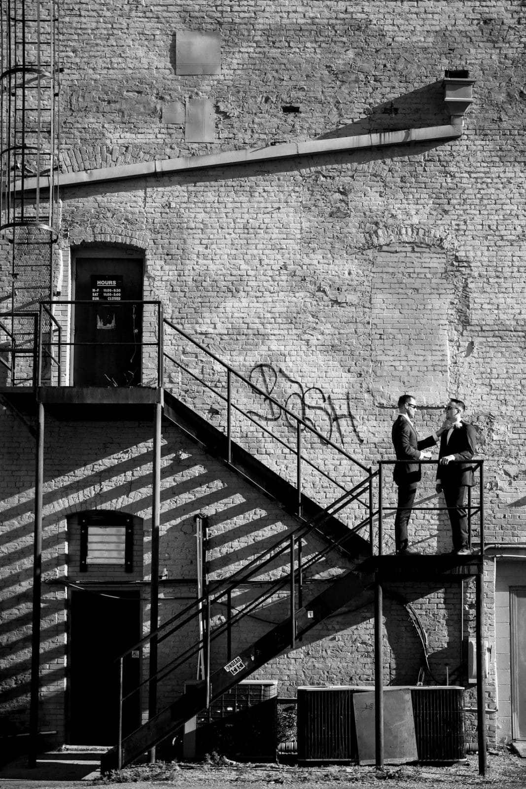Two people stand engaged in conversation on a metal staircase attached to a brick wall near Union Station. The wall, adorned with graffiti and a door marked 23406, casts intricate shadows from the railing, creating an urban and industrial setting perfect for unique Raleigh wedding photography.