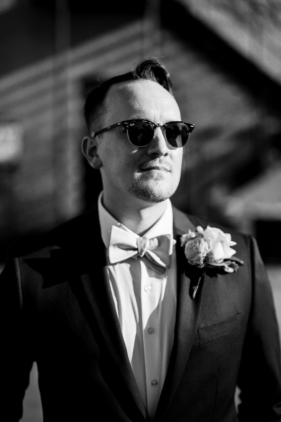 A man in a suit wearing sunglasses looks confidently into the distance, embodying the elegance captured in Union Station Raleigh wedding photography. He sports a bow tie and a flower boutonniere on his lapel. The black-and-white image features a softly blurred background, adding timeless charm.