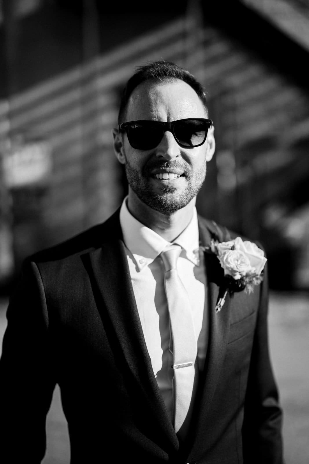 A man wearing sunglasses and a suit with a boutonniere is smiling in an outdoor setting at Union Station Raleigh. The image is in black and white, creating a classic, timeless feel, perfect for wedding photography.