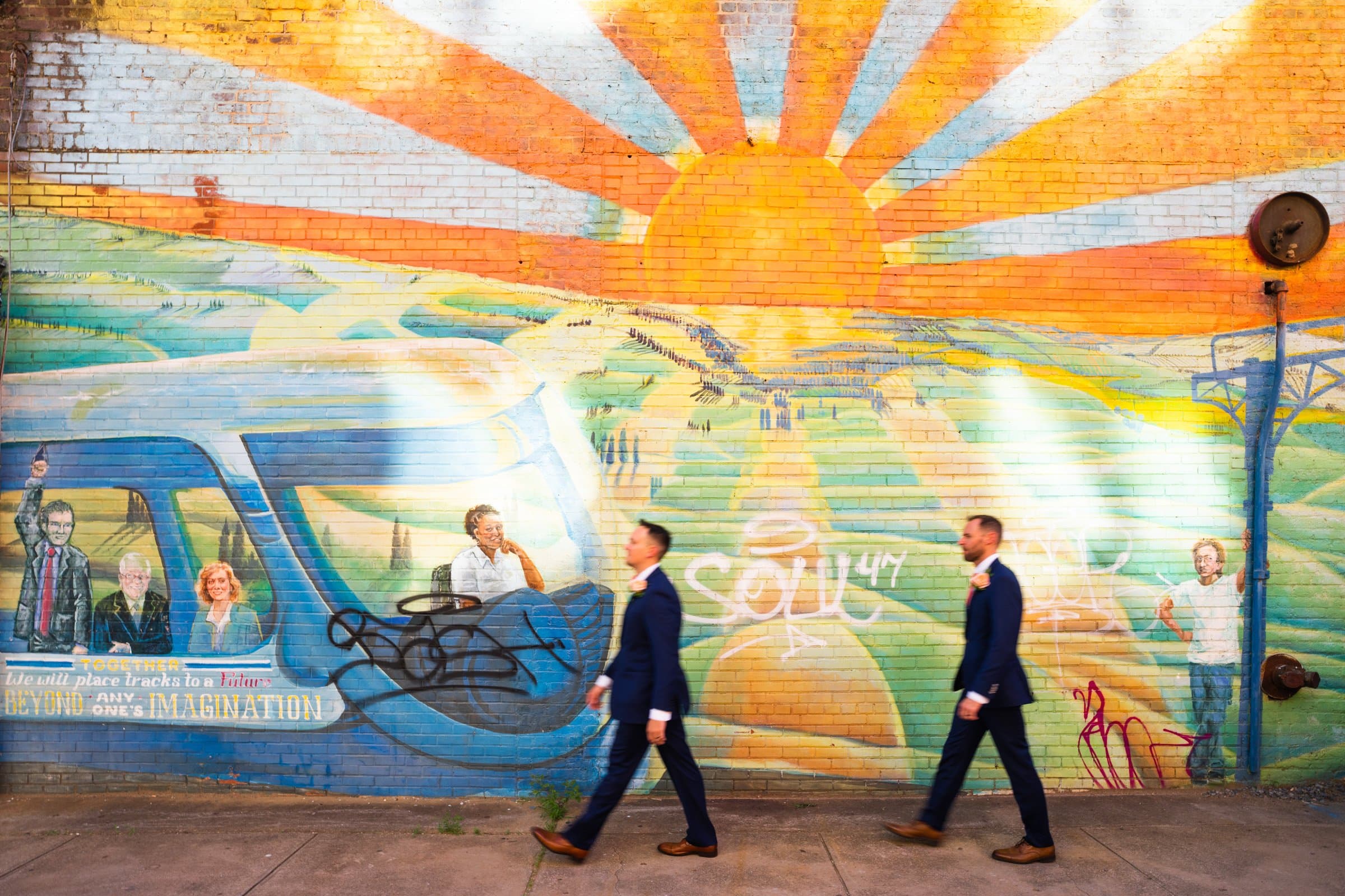 Two people in suits walk past a vibrant mural near Union Station in Raleigh, depicting a sunburst over hills and people in a colorful landscape. The mural includes a van with artistic text and graffiti-style elements, creating an ideal backdrop for wedding photography.