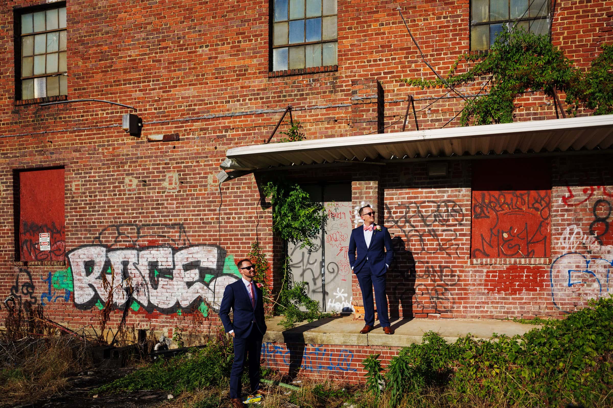 Two people in suits stand outside a graffiti-covered brick building, capturing a unique Raleigh wedding photography moment. One is on the ground while the other stands on a small platform. The scene is illuminated by bright sunlight, casting playful shadows reminiscent of Union Station's charm.
