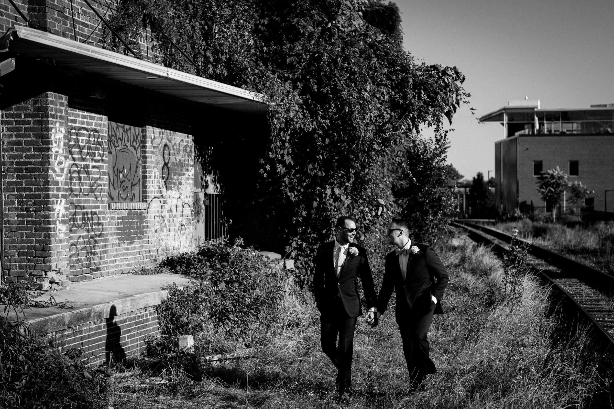 Two men in suits hold hands as they stroll past a graffiti-covered brick building near Union Station Raleigh. The black and white image captures a casual, intimate moment beside the train tracks, blending urban charm with wedding photography's timeless grace amidst overgrown grass.