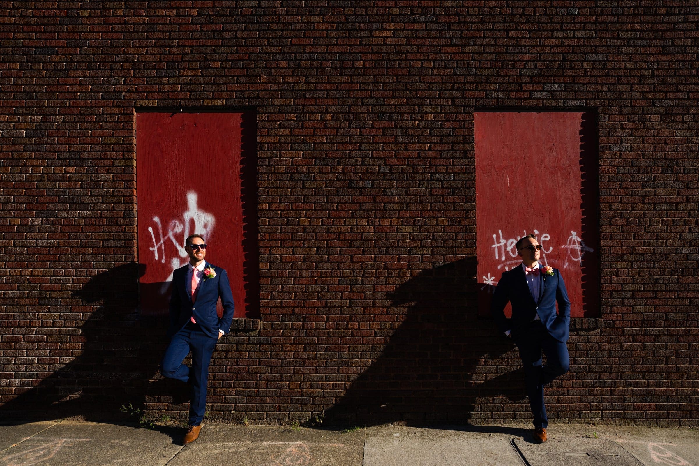 Two men in matching blue suits with pink bow ties stand confidently against a brick wall with red accents and graffiti. Their shadows are cast on the wall in bright sunlight, evoking the vibrant essence of Union Station Raleigh wedding photography.