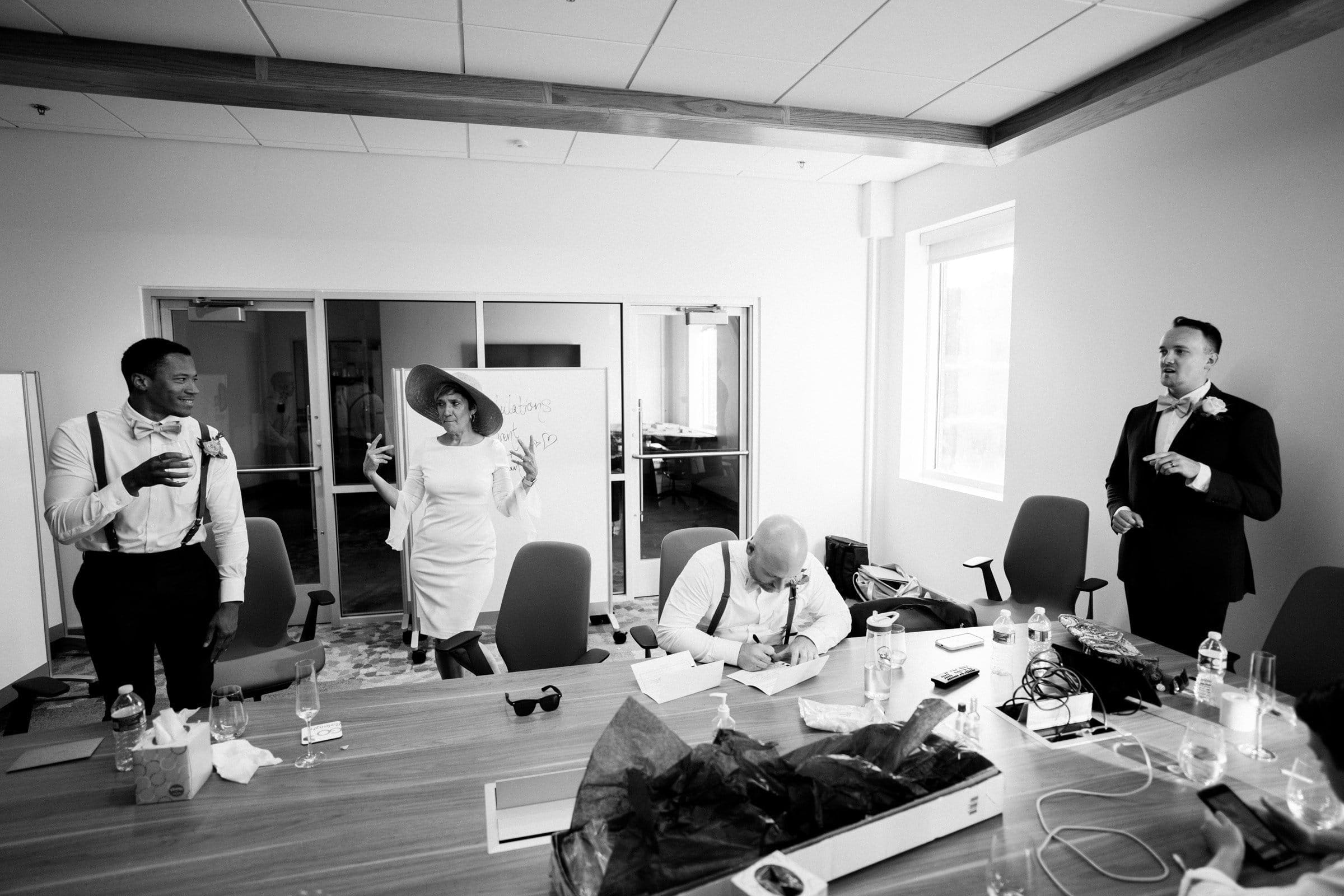A black and white photo captures the lively spirit of a Union Station Raleigh event. A person in a white dress and hat gestures playfully, while others in suits and someone writing at a cluttered table smile and chat, embodying the joyful essence of wedding photography.