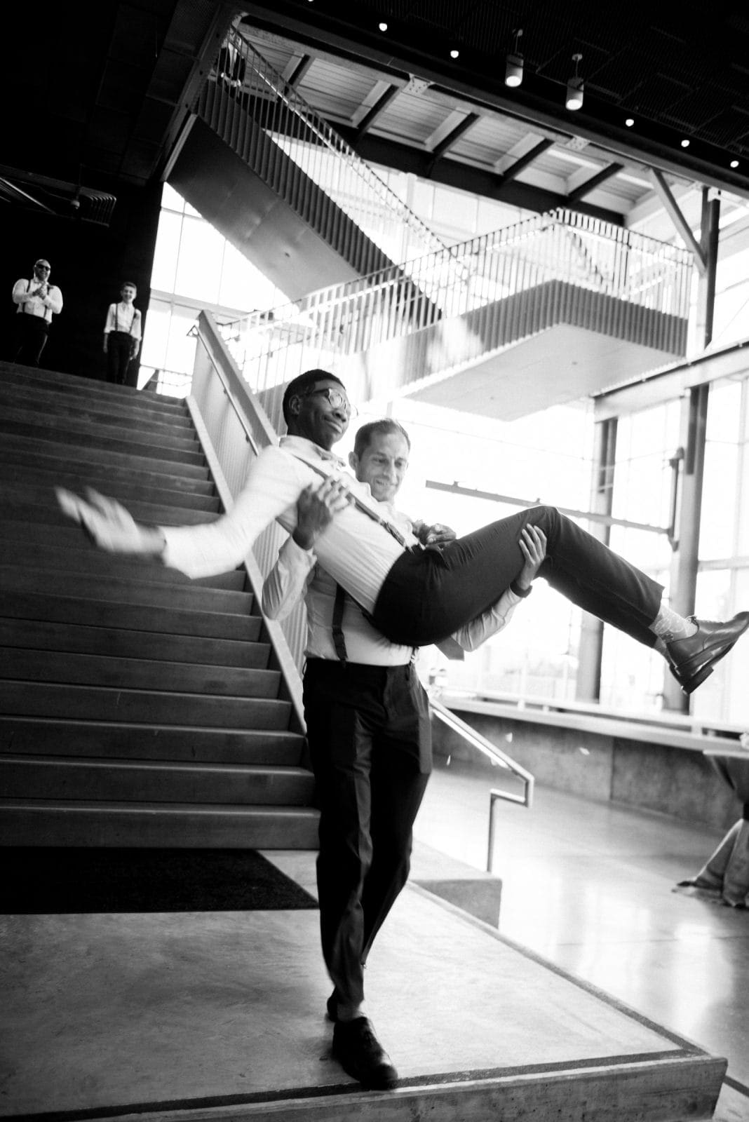 A black-and-white photo captures a man playfully carrying another in a light-hearted manner inside Raleigh's Union Station. With large windows and a staircase, the modern building provides a spacious backdrop, embodying the fun spirit often sought in wedding photography.