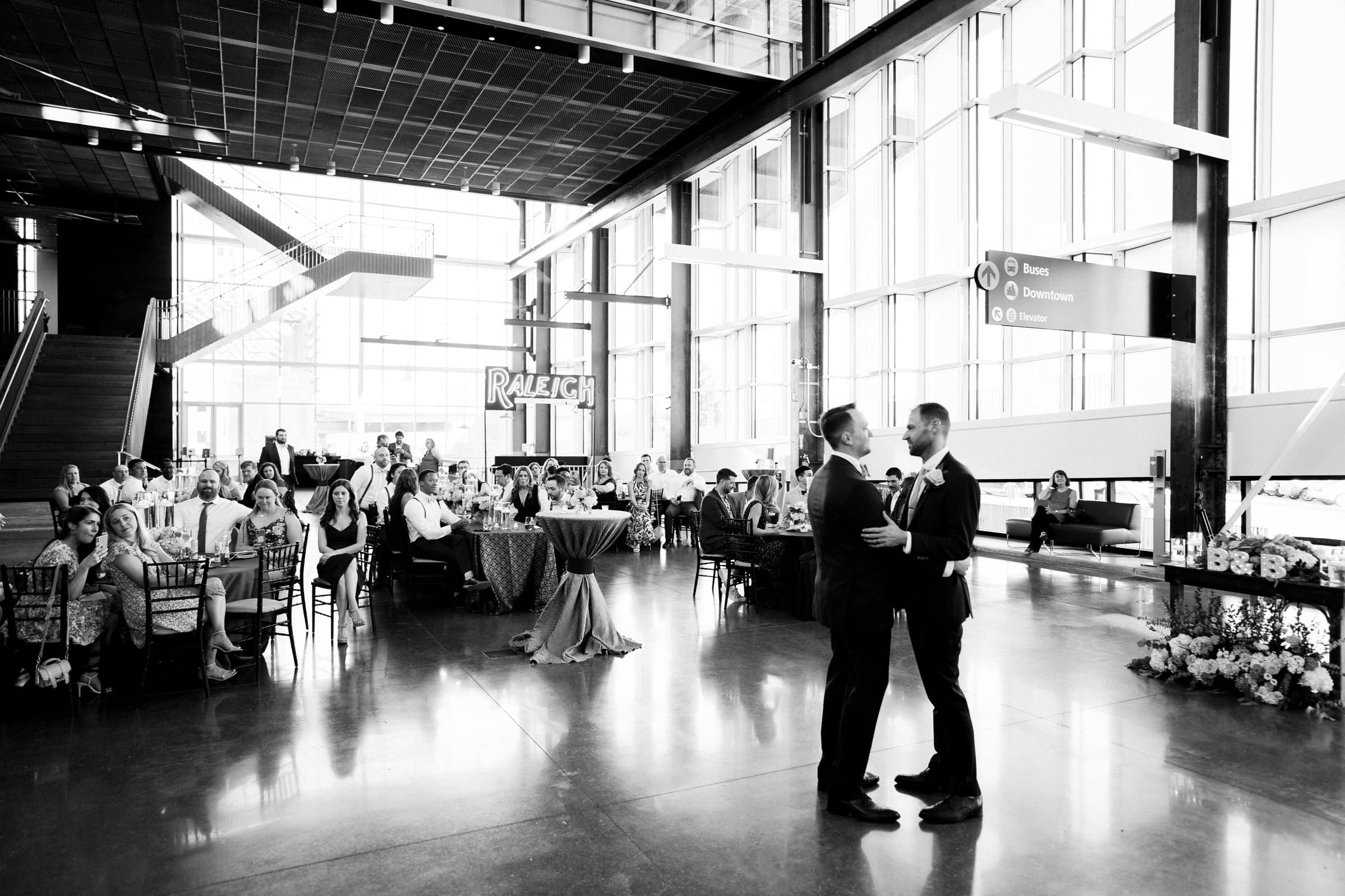 A black-and-white photo captures two men facing each other on a dance floor, surrounded by seated guests at a wedding reception. The modern architecture and large windows suggest an elegant venue like Union Station Raleigh, perfect for timeless wedding photography.
