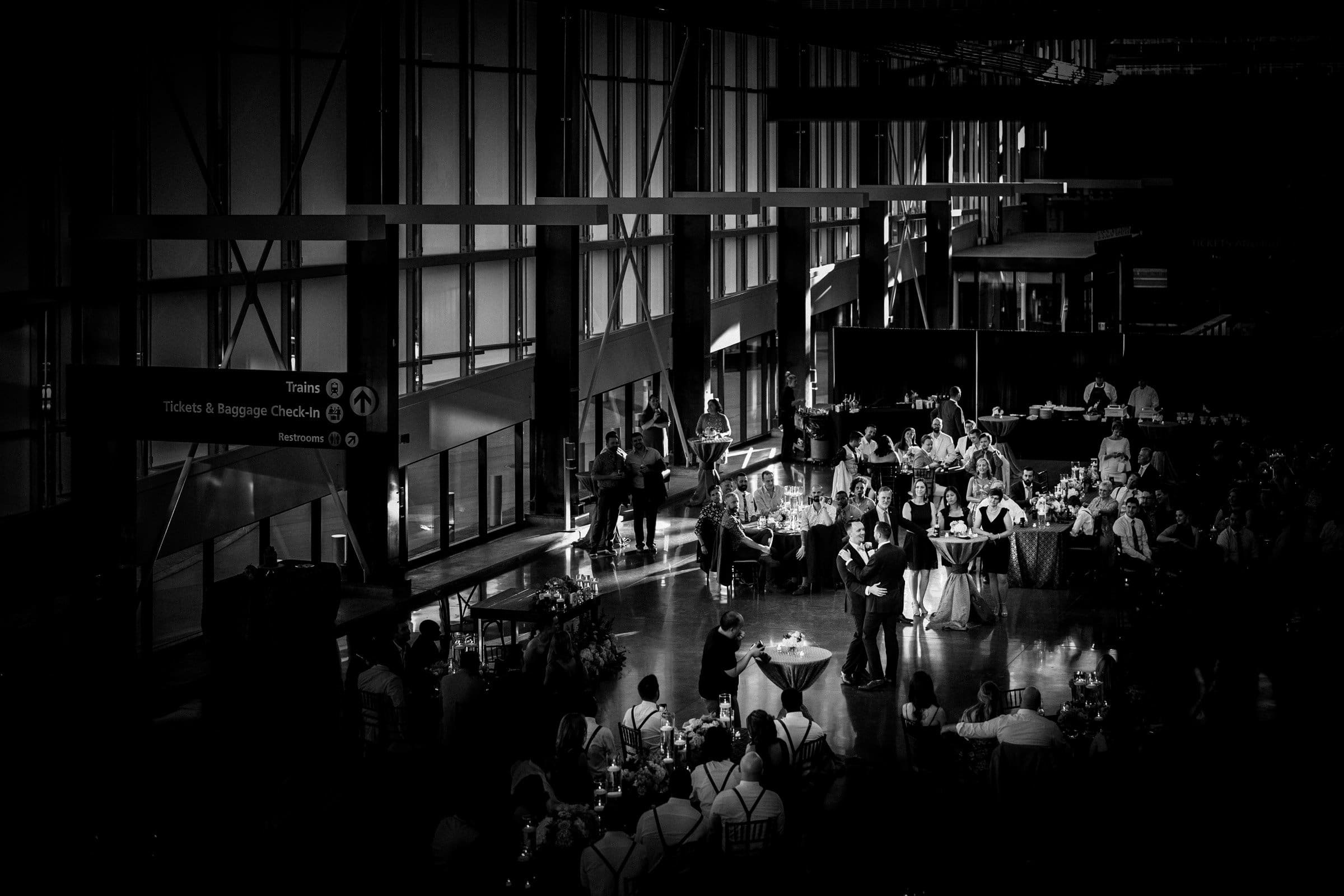 A black and white photo captures a wedding reception in a large hall, likely Union Station. Guests at tables watch a couple in the center holding a microphone. Light streams through tall windows, casting shadows on the floor, while signs for trains and baggage check add charm to the Raleigh scene.
