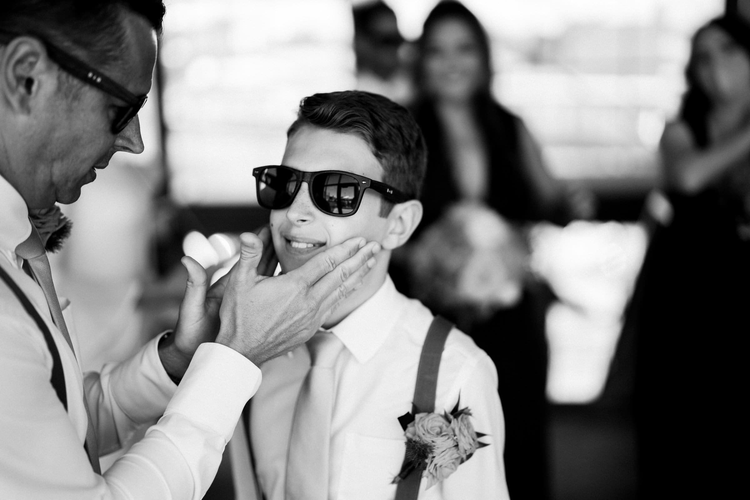 A black and white photo captures a man in sunglasses gently cupping the chin of a boy, both dressed formally with the boy sporting suspenders and a flower on his shirt. They seem to be at a Union Station Raleigh wedding, with blurred guests mingling in the background.