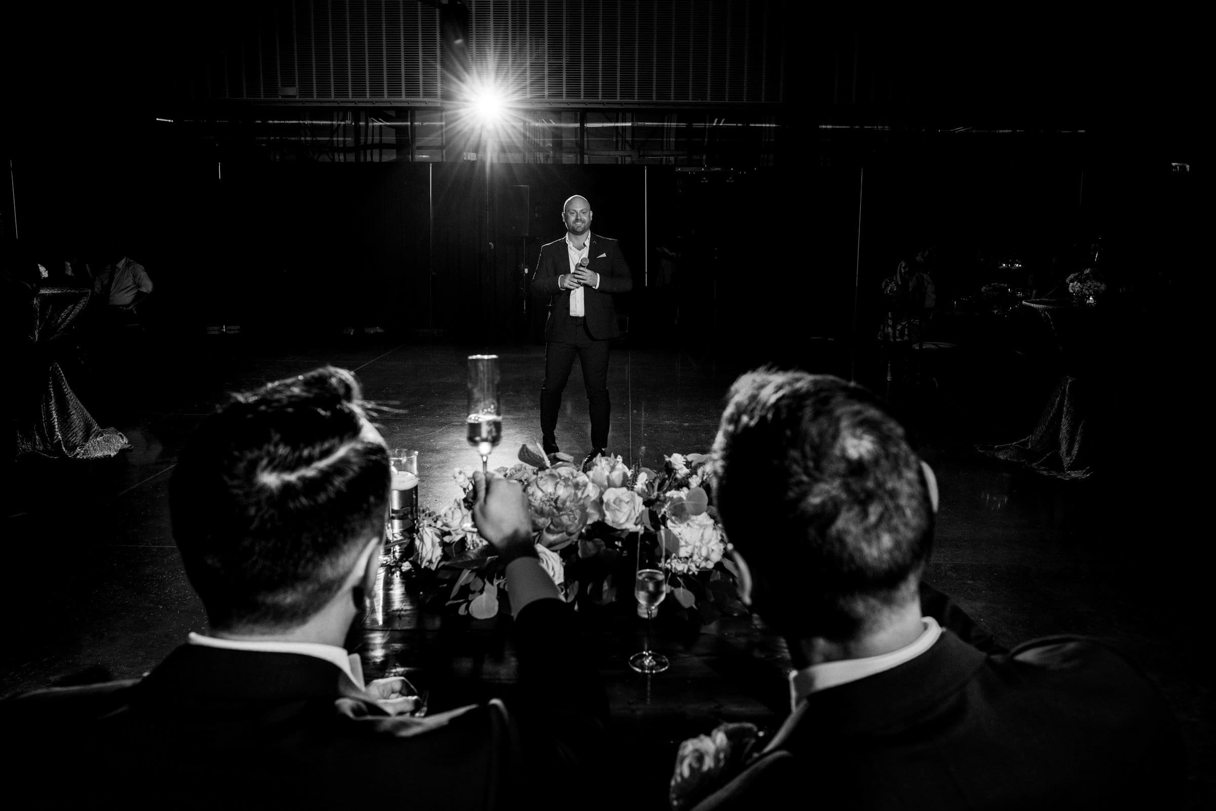 A man in a suit stands under a spotlight, holding a microphone, speaking to a seated audience in the dimly lit room of Union Station. Two people with drinks in hand listen attentively while the floral arrangement, perfect for wedding photography, decorates the table in the foreground.