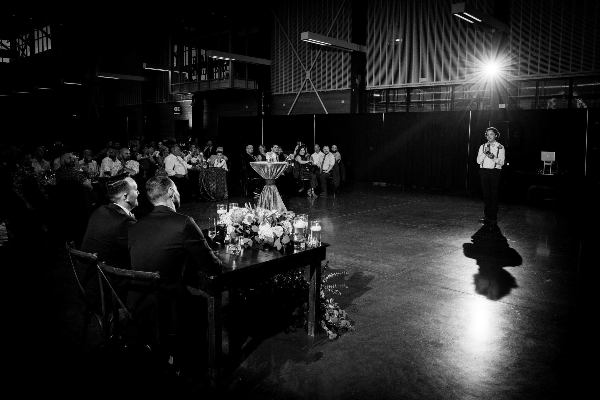 A black and white photo captures the magic of a Union Station Raleigh wedding reception. A person with a microphone stands on the right, addressing the audience seated at tables adorned with floral arrangements and candles. A bright light shines from the back, creating an enchanting ambiance.