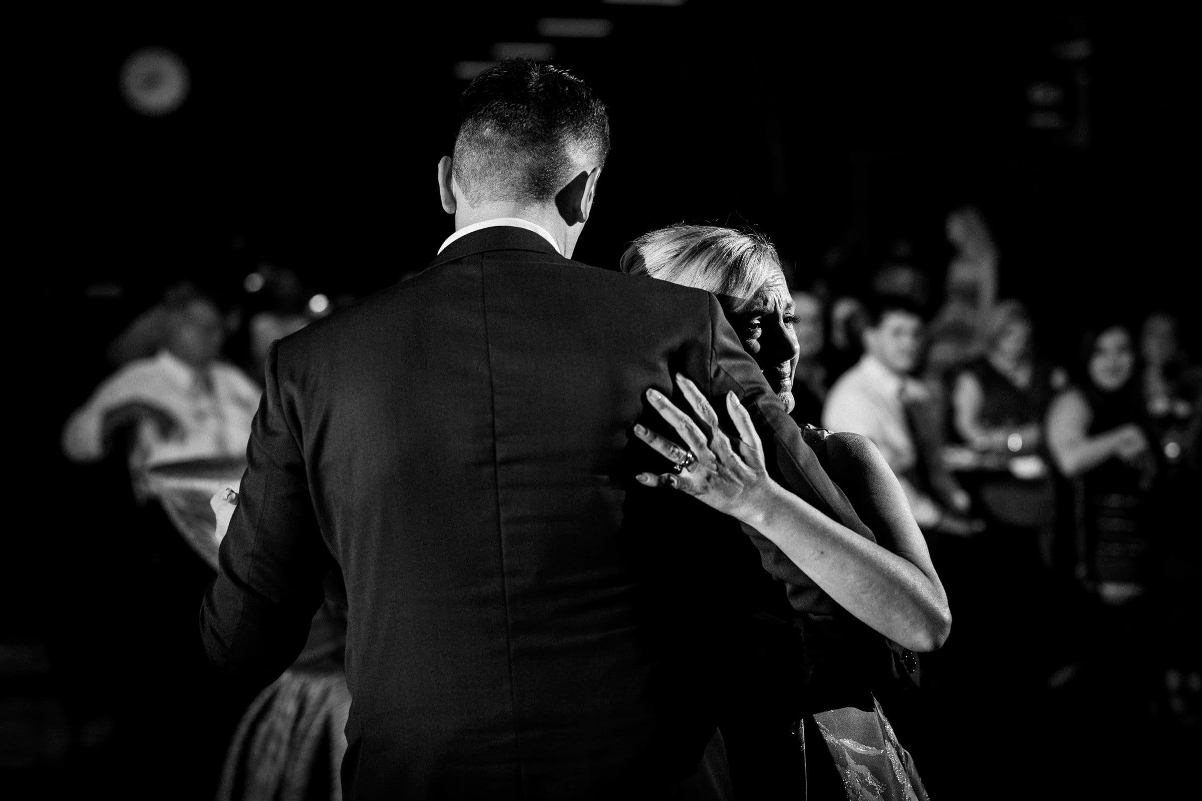 A black and white image captures a couple dancing closely in a dimly lit room. The woman, wearing a dress, has her hand on the man's shoulder, highlighting a ring on her finger. In the background, seated guests at Union Station Raleigh watch them, an intimate moment in wedding photography history.