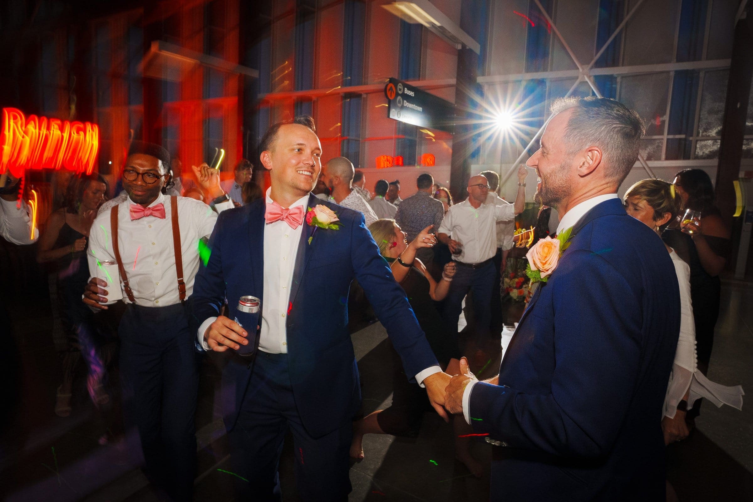 A festive party scene unfolds with guests dancing joyfully. Two men in suits, each sporting pink bow ties, hold hands and smile warmly at each other. The lively atmosphere, captured like Raleigh wedding photography at Union Station, features colorful lights and a bright flash illuminating the celebration.