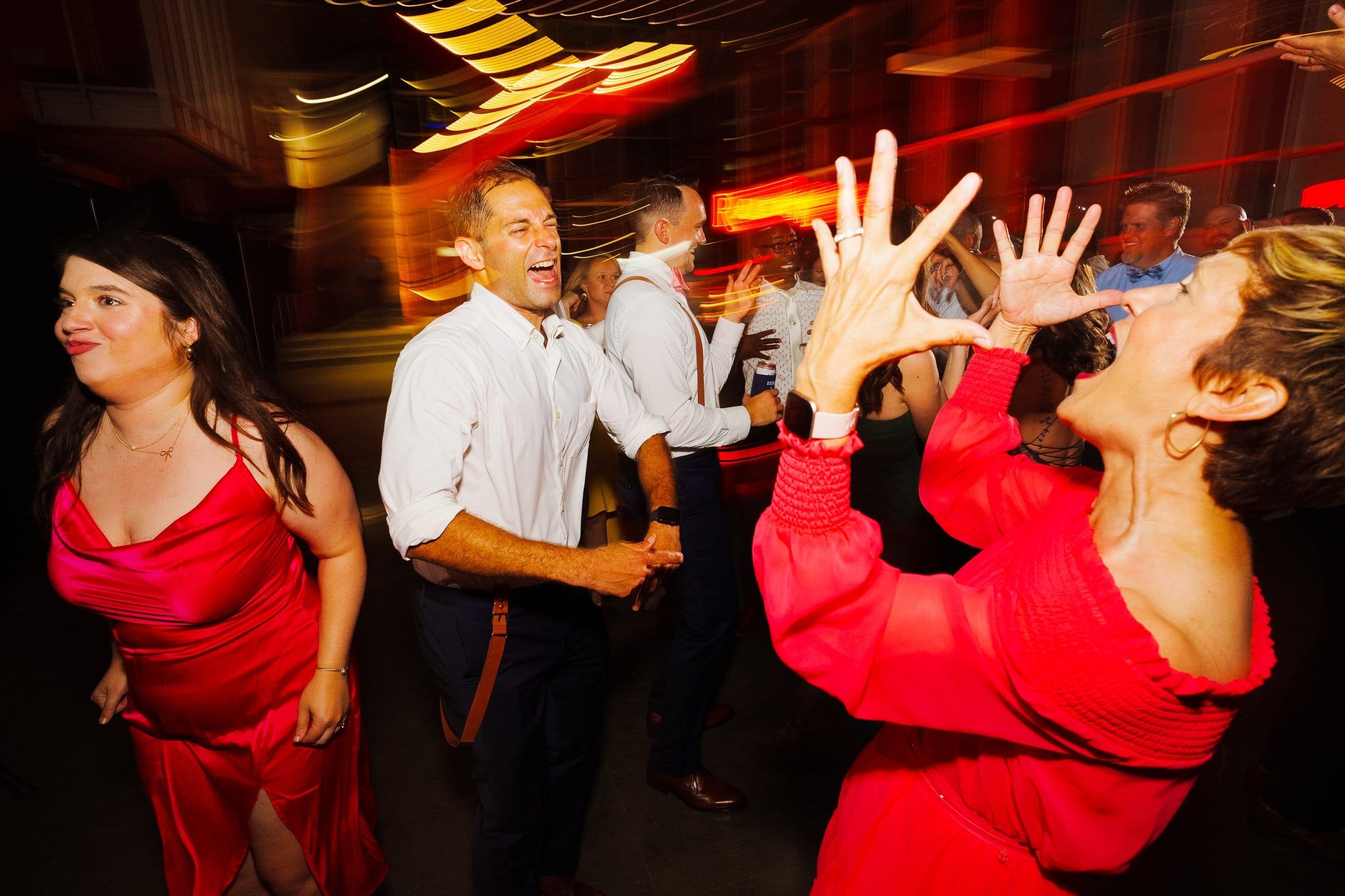 People joyfully dancing in a lively, colorful setting at Union Station Raleigh. A woman in the foreground enthusiastically gestures with her hands, wearing a red dress. Others around her also wear bright clothing, and the background features vibrant lights and motion blur—perfect for wedding photography.