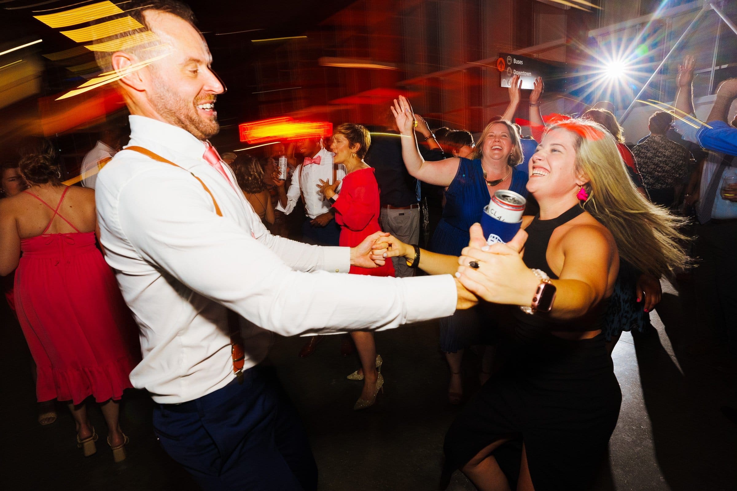 A man and woman smiling and dancing energetically at a lively event. The woman, in a black dress, holds a can, while the man sports suspenders with a white shirt. Captured in classic Union Station Raleigh wedding photography style, the blurred background reveals others enjoying the occasion.