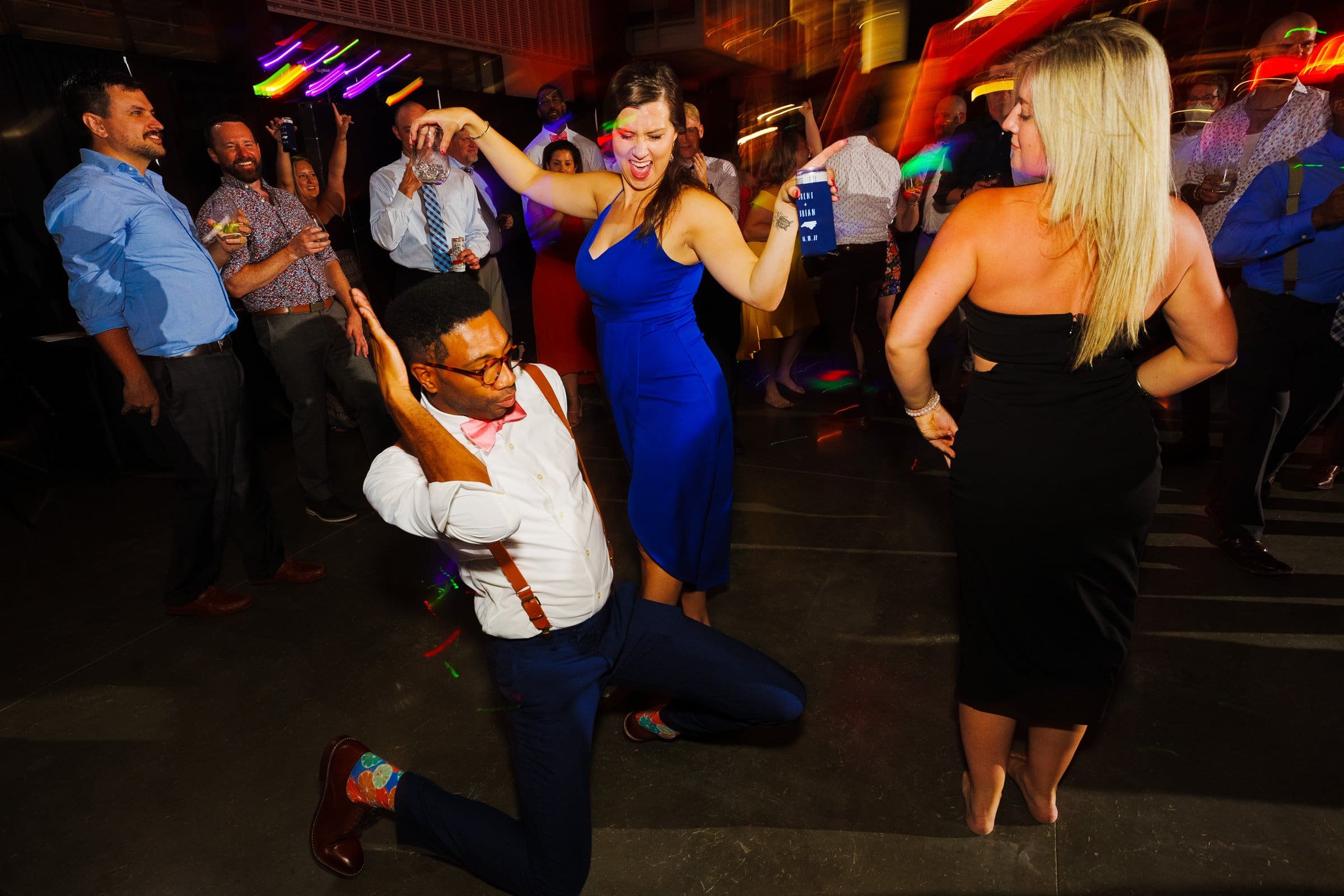 A lively party scene reminiscent of a Union Station wedding with people dancing energetically on a dimly lit dance floor. One person kneels playfully, striking a pose in colorful socks, while others enjoy the music and atmosphere, drinks in hand. Bright lights amplify the festive mood.