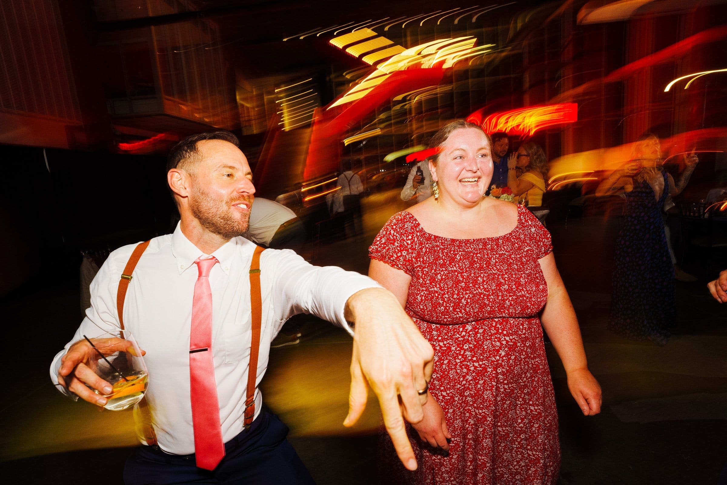 A man and woman joyfully dance at a party, captured perfectly in the style of Union Station Raleigh wedding photography. The man sports a white shirt with a pink tie and suspenders, drink in hand, while the woman dazzles in a red floral dress against the backdrop of vibrant, blurred lights.
