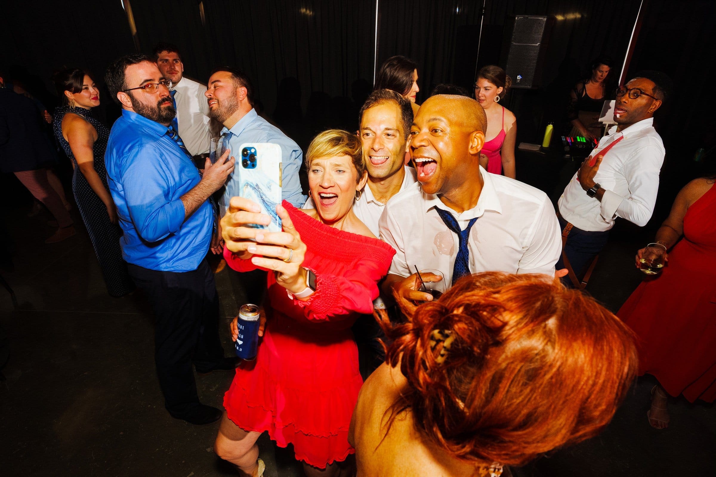 At a lively party at Union Station Raleigh, a group is gathered, smiling and taking a selfie. Dressed in festive attire, they seem to be enjoying themselves with others dancing and socializing in the background—a perfect moment for wedding photography.