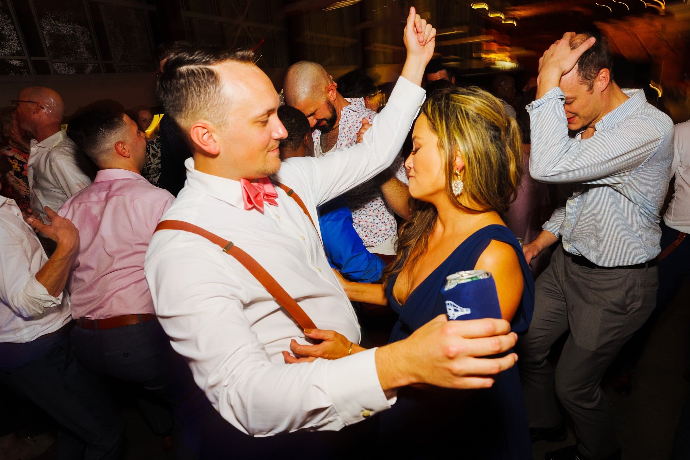 A lively scene unfolds at a party reminiscent of a Union Station Raleigh wedding. People dance energetically; a man in a white shirt and bow tie whirls with a woman in a blue dress. The dimly lit room buzzes with vibrant joy, the blurred motion capturing every exciting moment of the celebration.