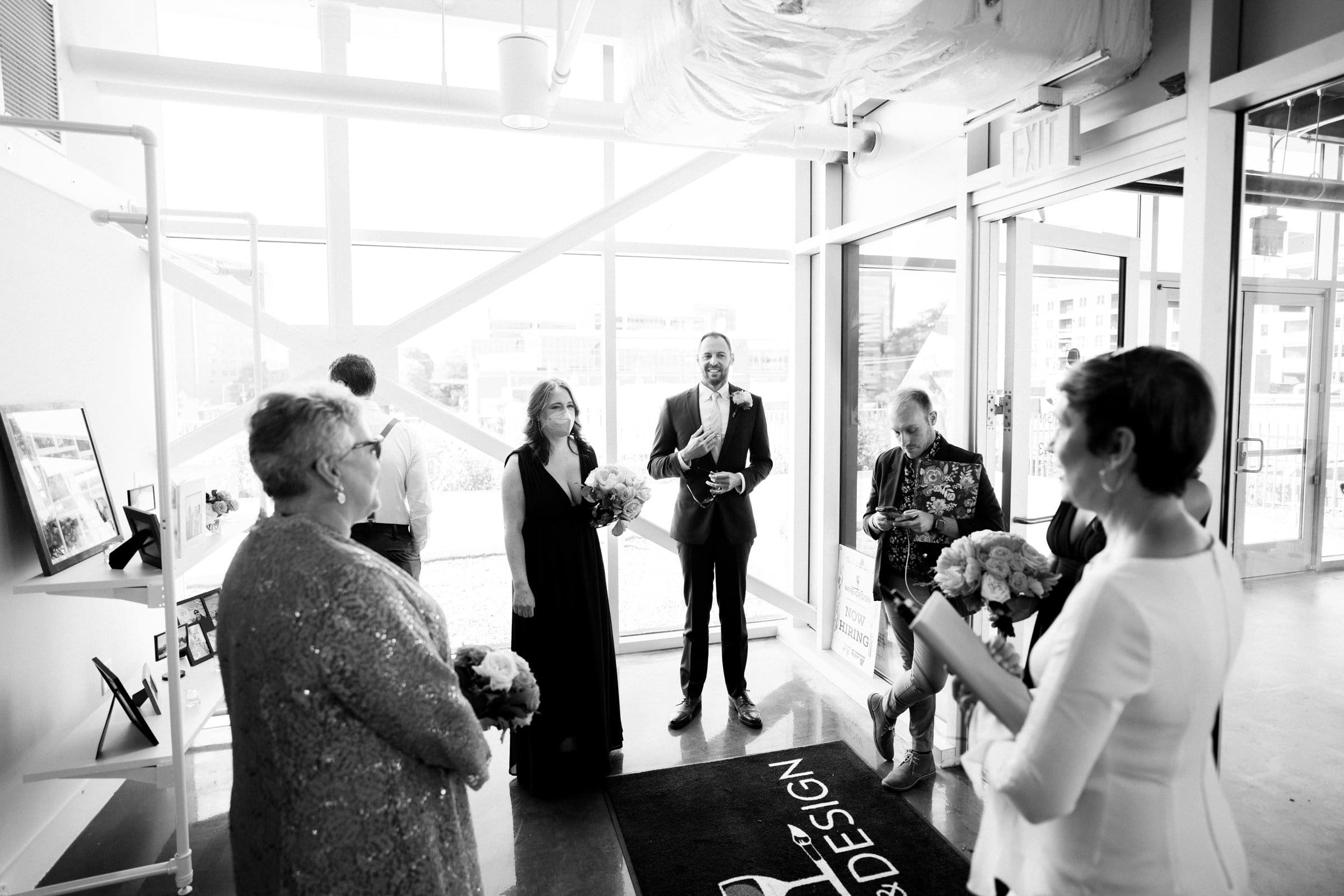 A black and white photo captures a small gathering in a modern indoor space reminiscent of Union Station. A man in a suit stands beside women holding bouquets, while another older man is nearby. People are visibly talking and smiling, with a large window revealing the Raleigh cityscape.