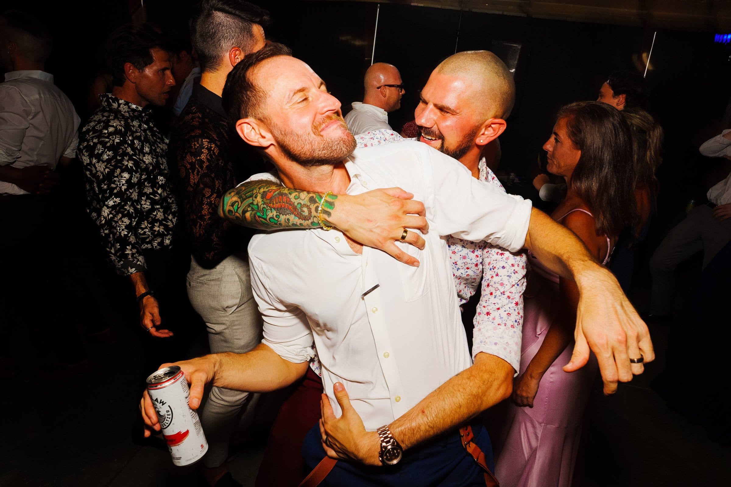 Two men in a lively dance embrace at a party, one holding a drink can. Both are smiling, surrounded by people dancing in the background. Capturing this joyful and energetic moment feels as vibrant as those shot during wedding photography at Union Station Raleigh.