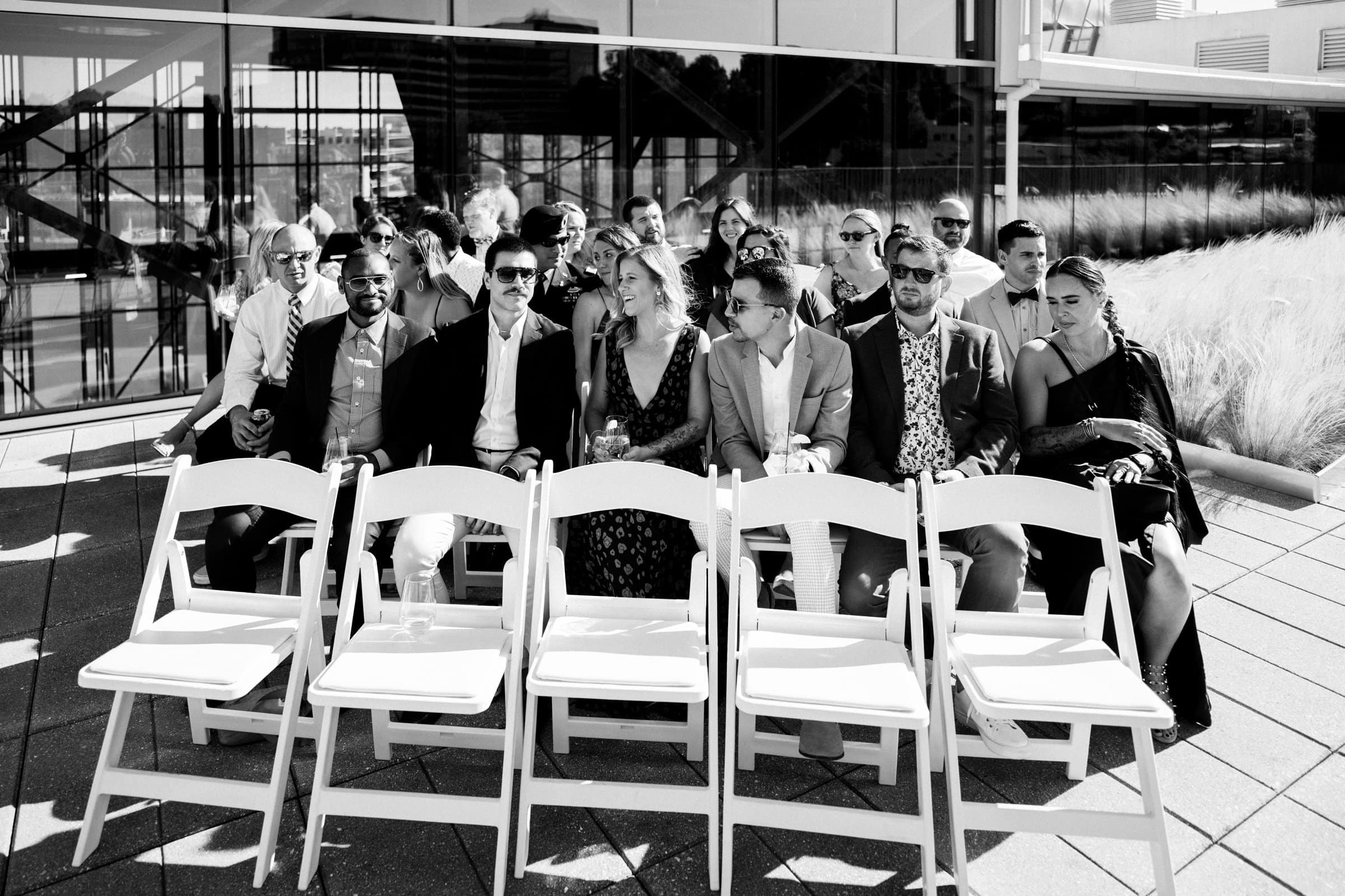 A group of people sits on white folding chairs arranged in rows outdoors, reminiscent of a wedding scene, in front of a modern building with glass windows. Captured in black and white like classic photography, sunlight casts shadows on the ground while some chairs remain empty.