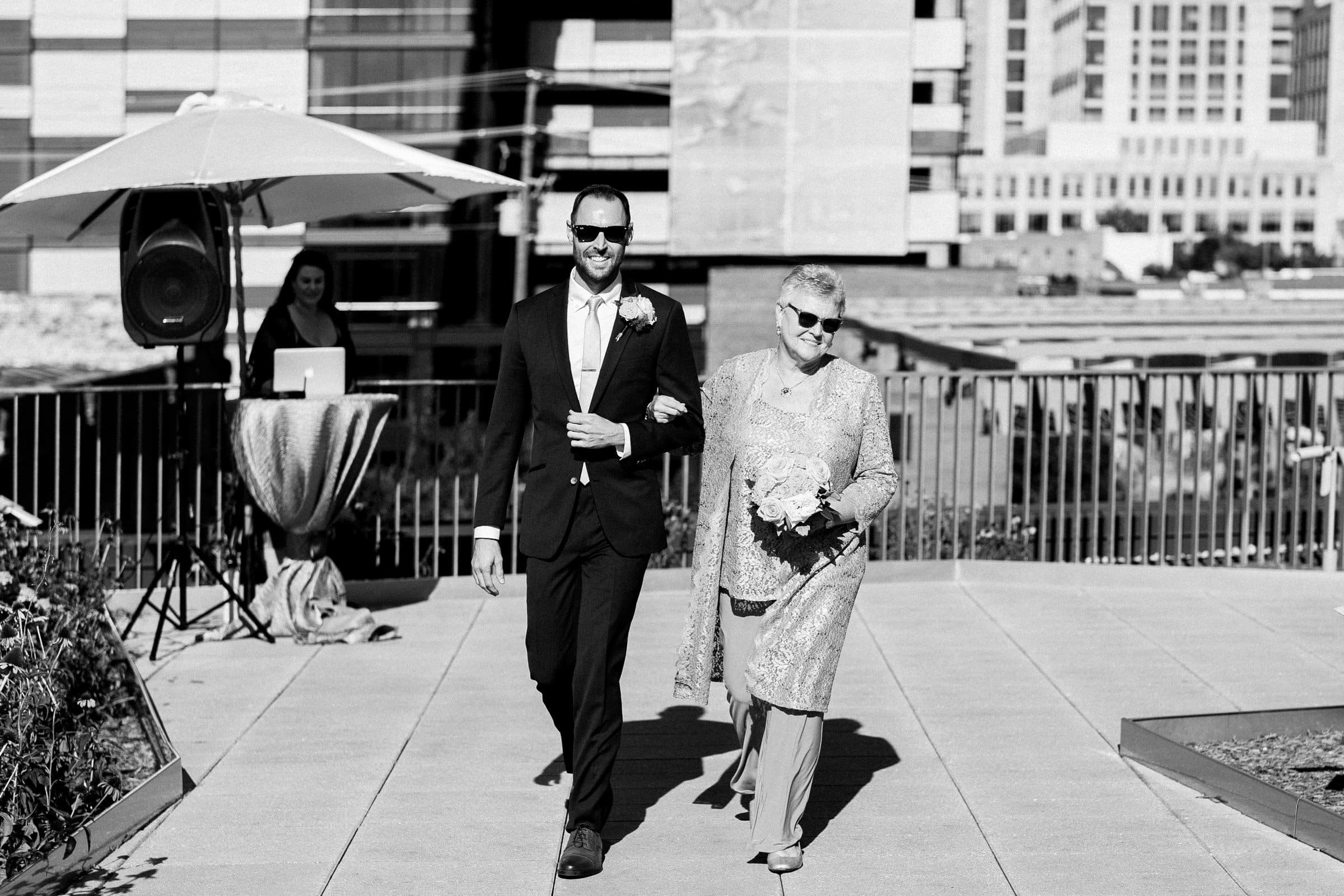 A man in a suit and sunglasses walks arm in arm with an older woman in a dress, holding flowers. They grace the terrace, city buildings framing their moment, reminiscent of elegant Union Station Raleigh wedding photography. Nearby, a DJ stands under an umbrella.