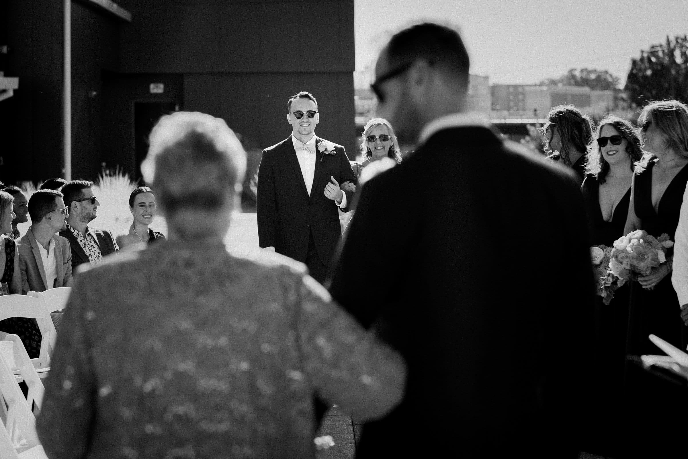 A black-and-white photo of a wedding ceremony at Union Station Raleigh. A person in a suit stands smiling in the center, surrounded by people seated and standing, some holding bouquets. The scene captures a celebratory moment outdoors, embodying exquisite wedding photography artistry.