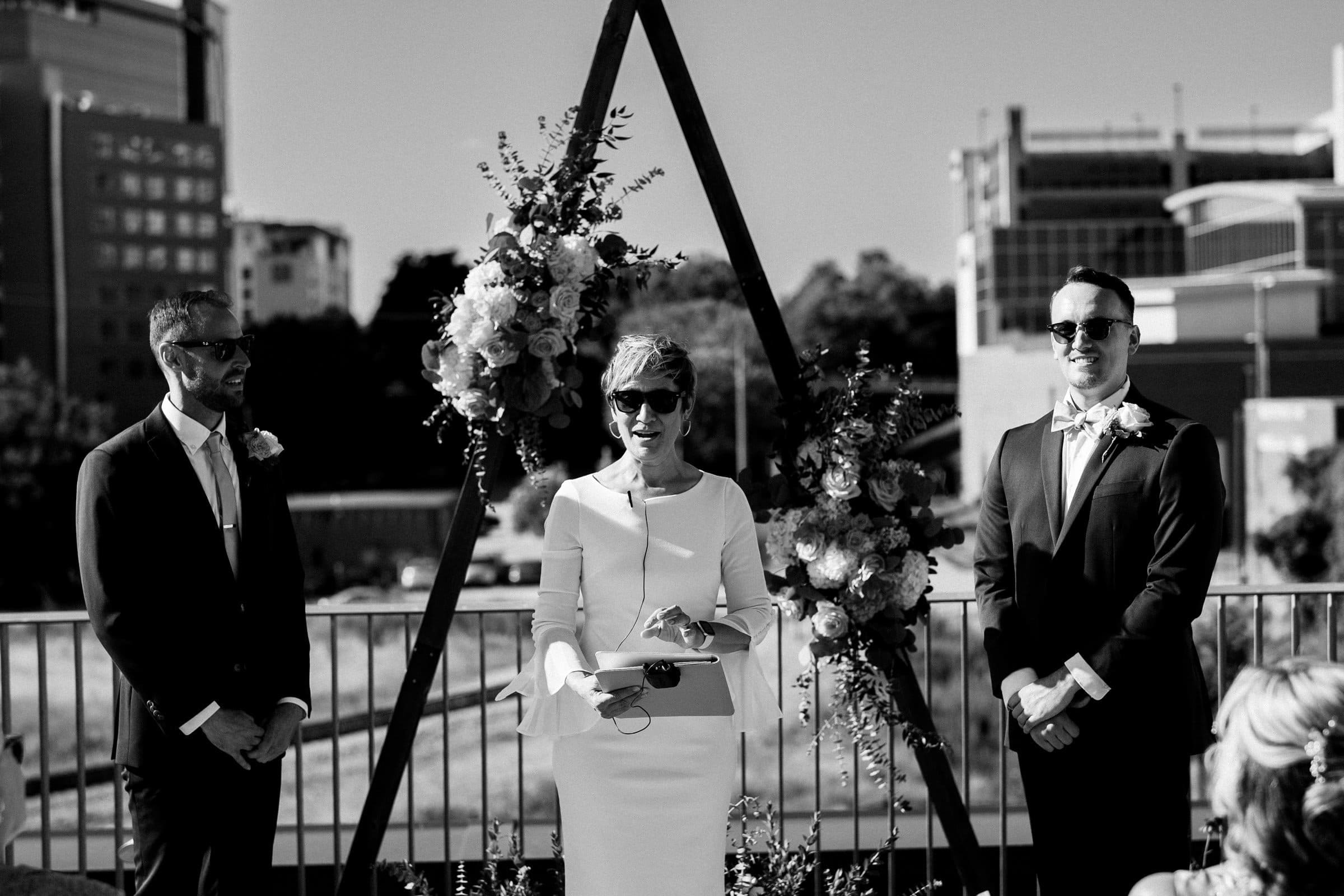 A black-and-white image captures a wedding ceremony at Union Station Raleigh. Two people in suits stand on either side of an officiant in a white dress, holding papers. A triangular arch adorned with flowers frames the scene, while urban buildings and trees provide a charming backdrop.