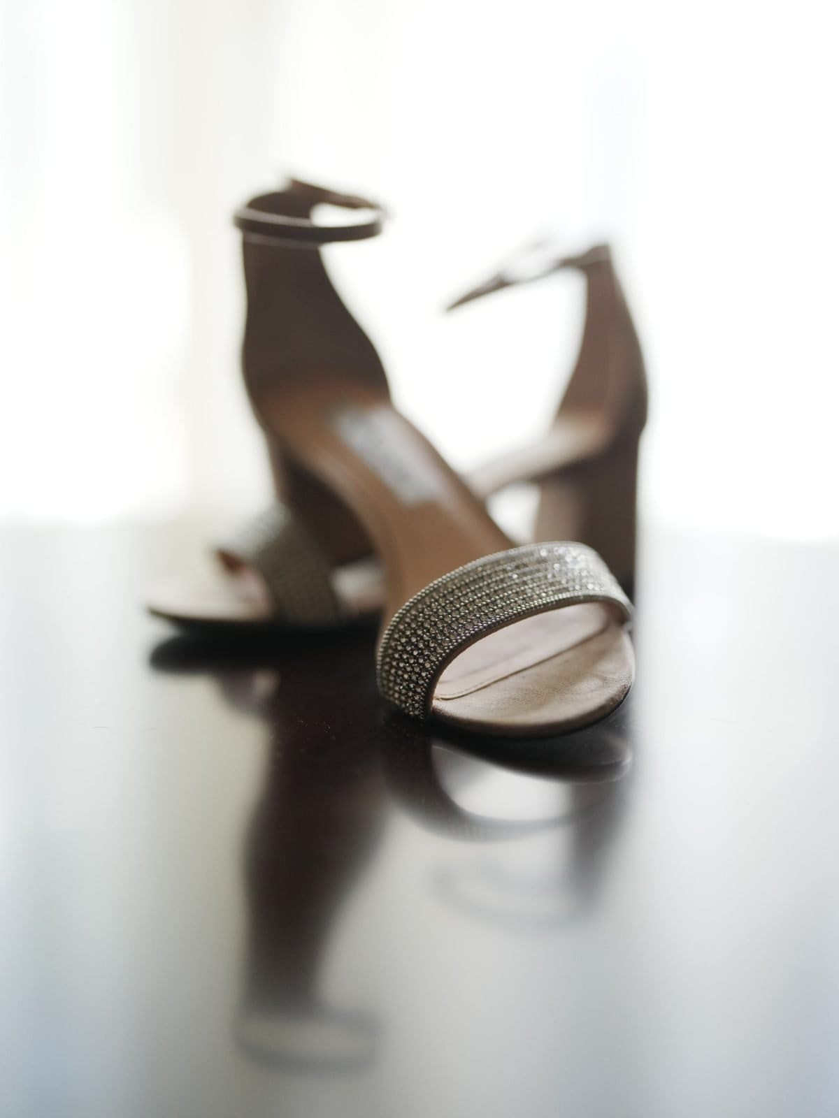 duke university wedding photography - sarah & case A pair of elegant, open-toed high heels with sparkling straps is displayed on a glossy surface, reminiscent of Duke University's wedding photography style. The background is softly blurred, highlighting the shoes as the focal point.
