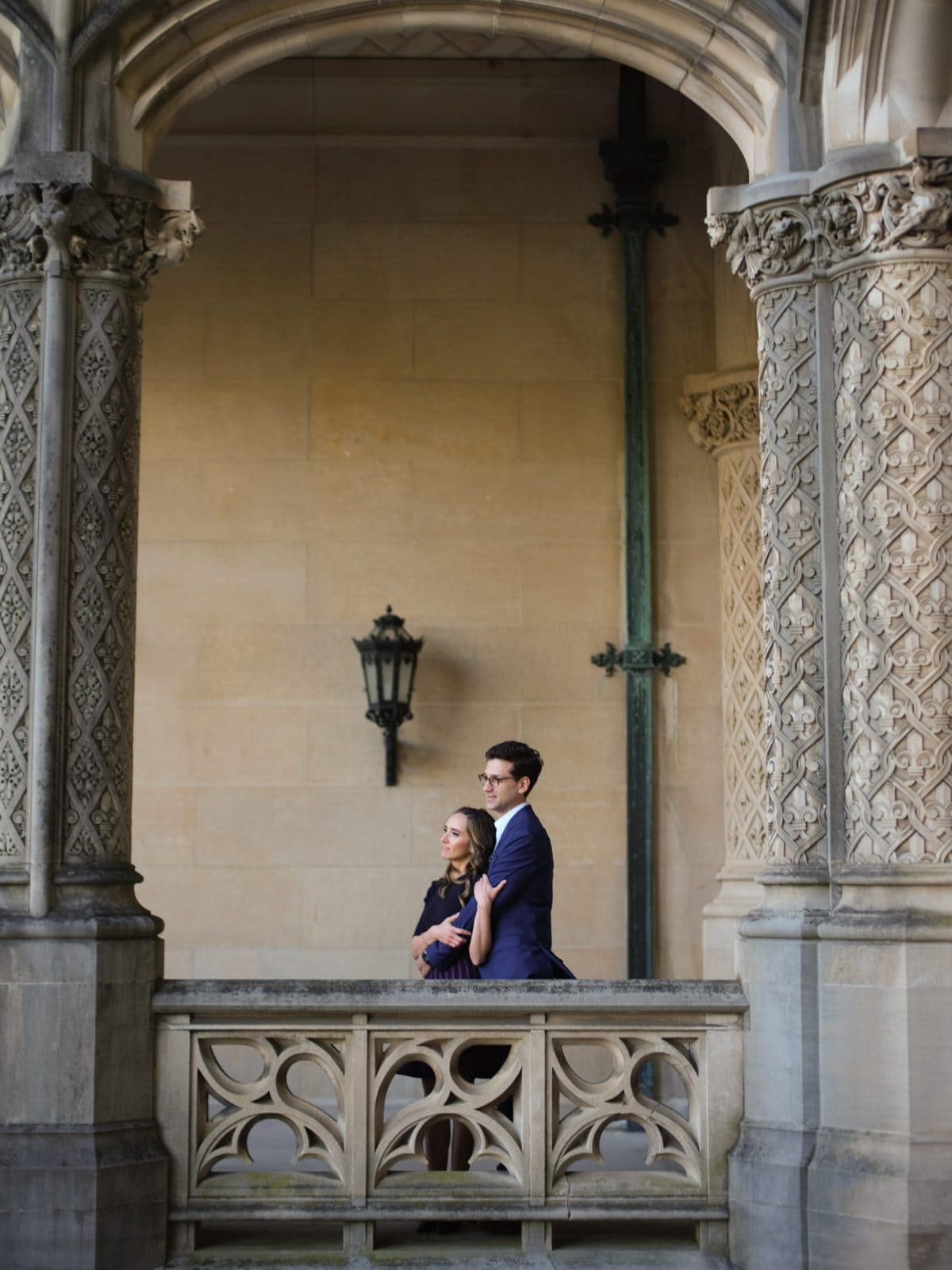 A couple stands on a stone balcony at the Biltmore Estate, surrounded by ornate columns and arches. The man, in a blue suit, has his arm around the woman in a light dress. A wall lantern and decorative carvings enhance the architectural elegance, perfect for engagement photography.