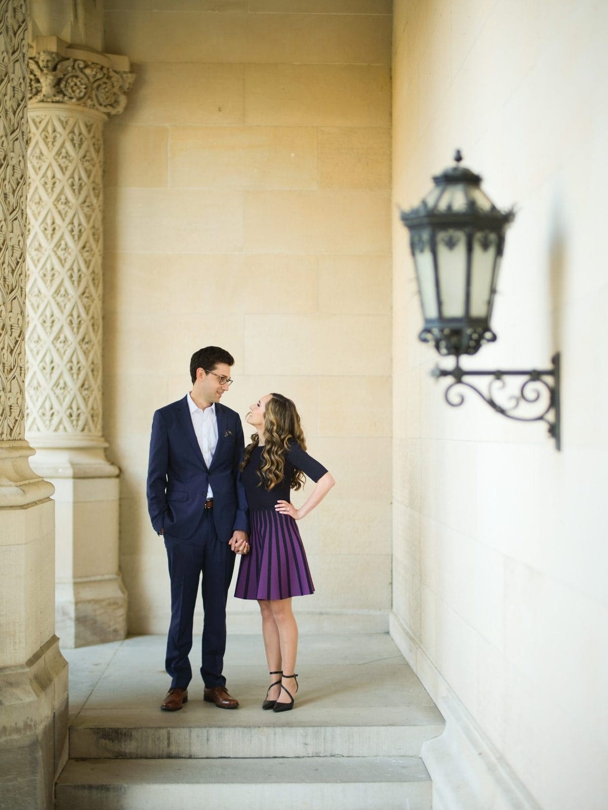 A man in a blue suit and a woman in a purple dress stand hand in hand in a corridor with ornate columns and a vintage lamp, embodying the charm of Biltmore Estate engagement photography. Their smiles create a warm, intimate atmosphere.