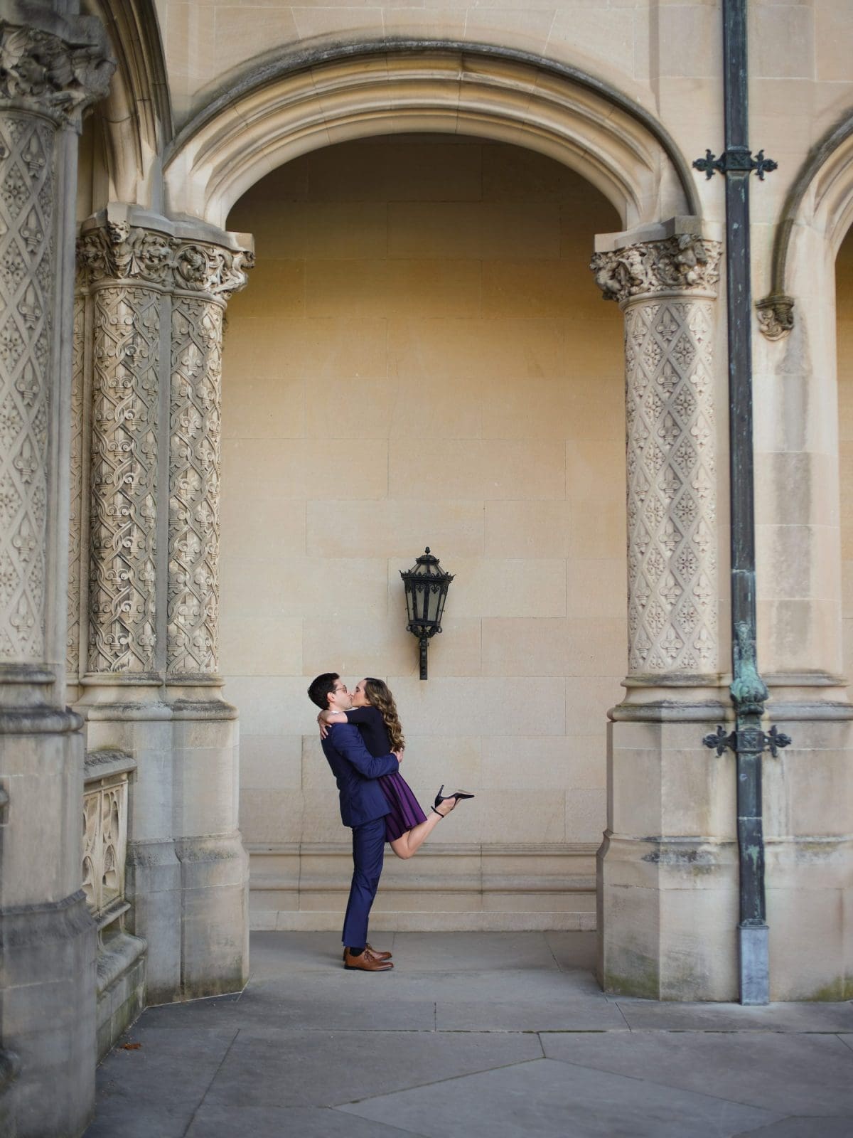 A couple embraces under the ornate stone arches of the Biltmore Estate. The man lifts the woman off the ground as they smile at each other, capturing a perfect engagement photography moment. She wears a dark dress and heels, while he is in a dark suit, framed by intricate carvings and a black lantern.