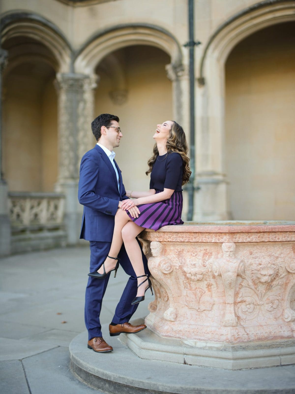 A man in a blue suit stands next to a woman sitting on a stone ledge at the Biltmore Estate. She's wearing a black top and purple-striped skirt, laughing with her head tilted back. The ornate architecture provides the perfect backdrop for their engagement photography session.