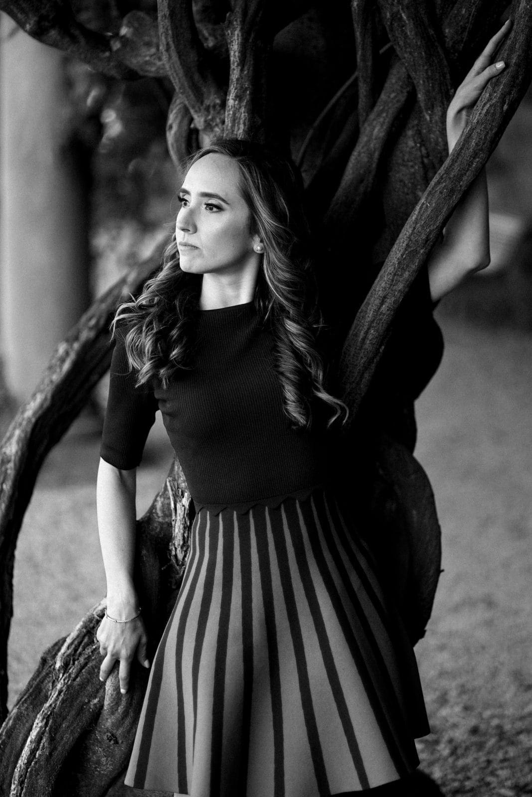A woman in a striped skirt and dark top leans against twisted tree branches at the Biltmore Estate, looking thoughtfully into the distance. The black and white image highlights her wavy hair and the textured branches, capturing an intimate moment in this engagement photography session.