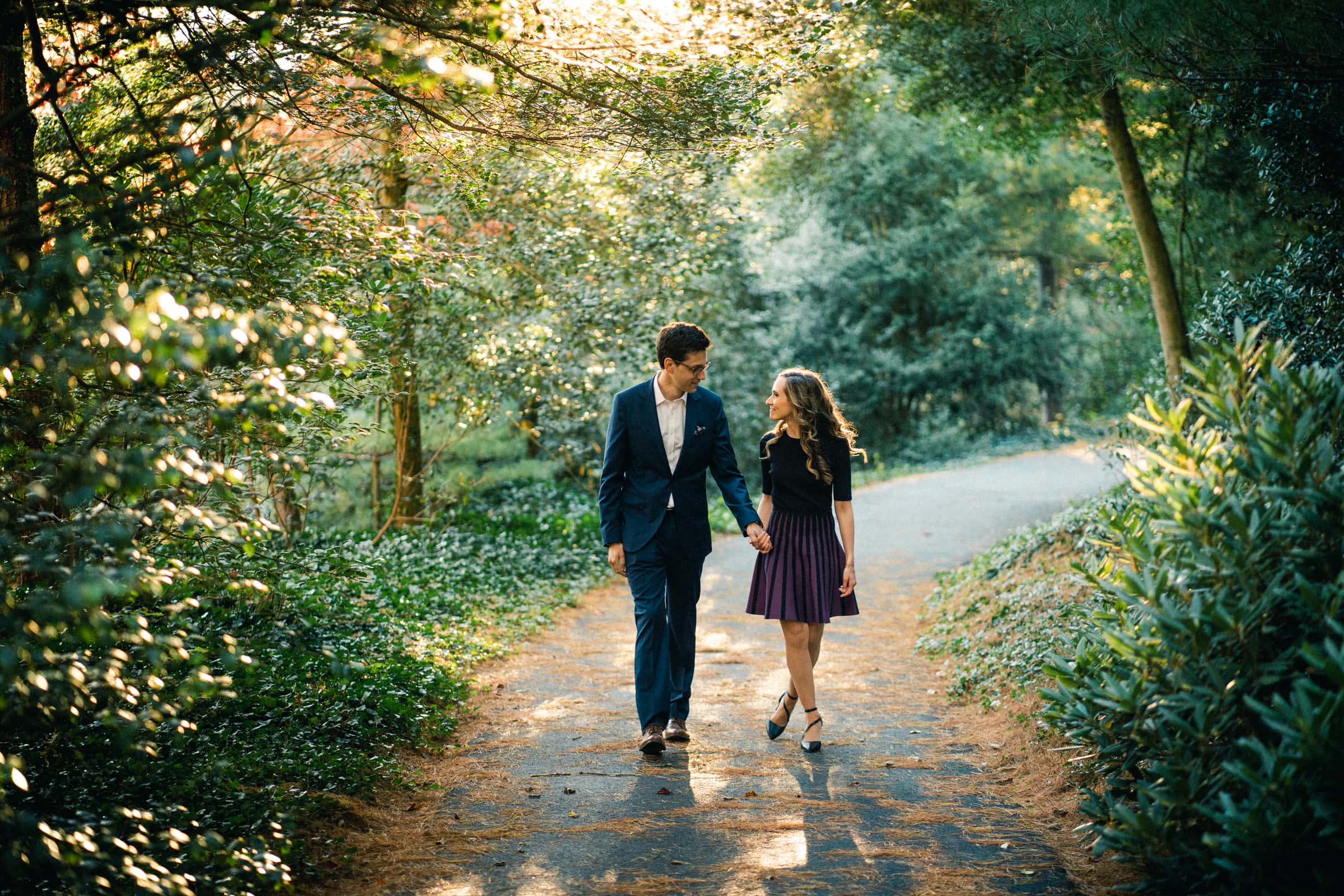 A couple holds hands, walking along a tree-lined path in a park reminiscent of the serene Biltmore Estate. The man is in a blue suit, while the woman wears a black top and purple skirt. Sunlight filters through the trees, capturing this perfect engagement photography moment.